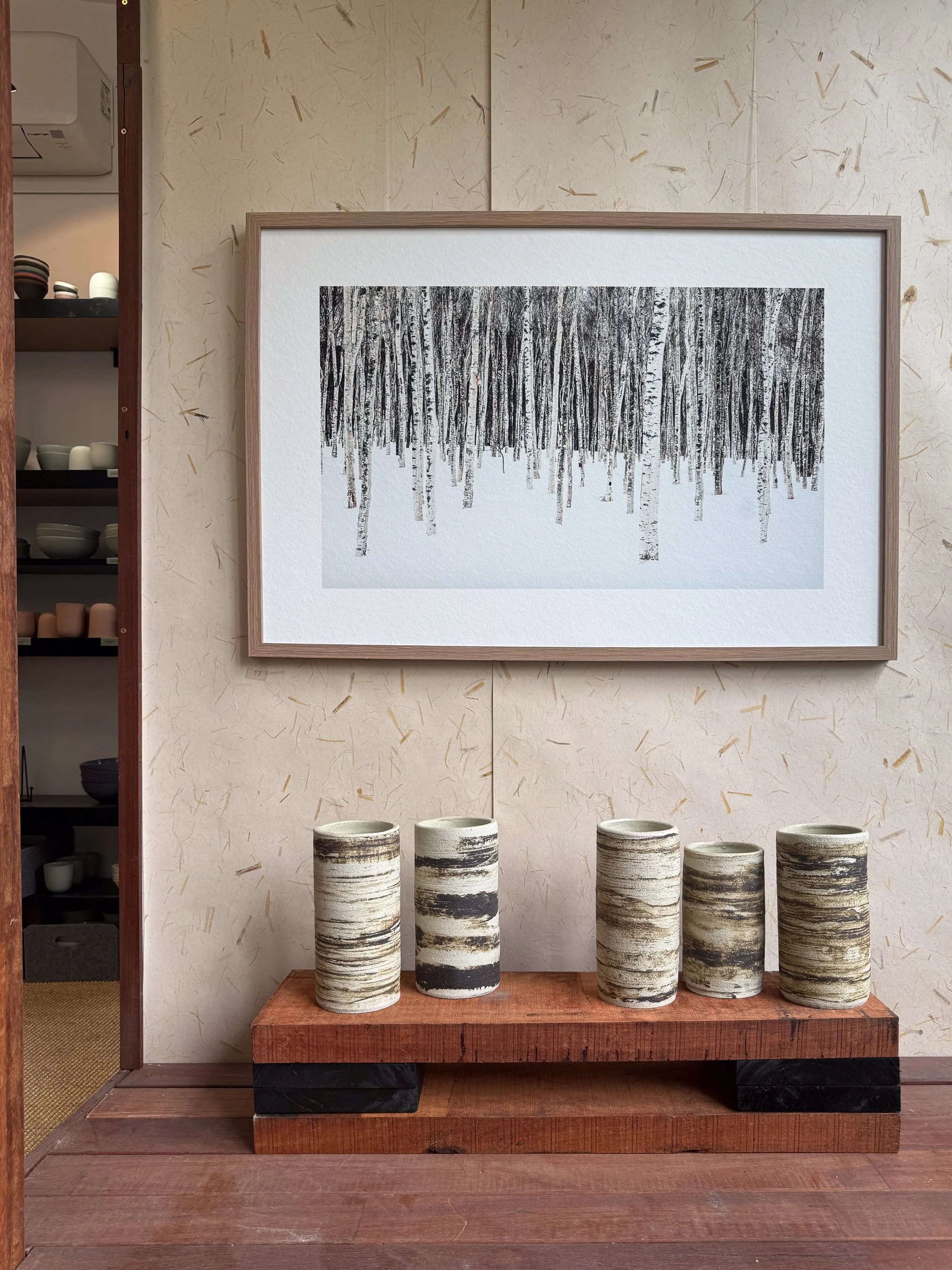 A wooden surface displays  striped ceramic vases of varying heights. An of a birch forest hangs on the wall behind the vases. The background wall has a textured beige pattern with scattered. To the left, open shelves with bowls and cups are visible.