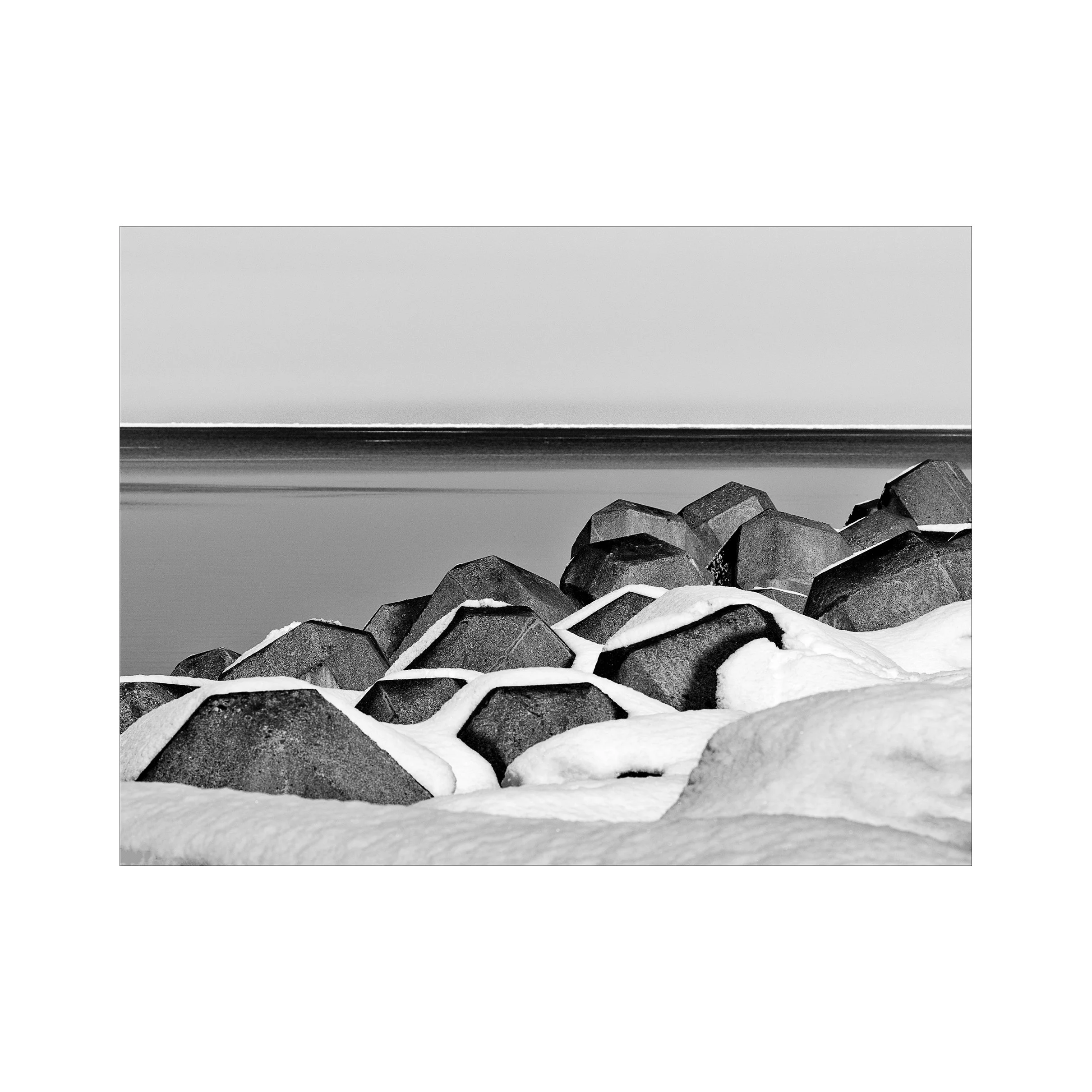Black and white photograph of large rocks or concrete blocks on snow-covered ground near a body of water,a shoreline with a blurred horizon in the background.