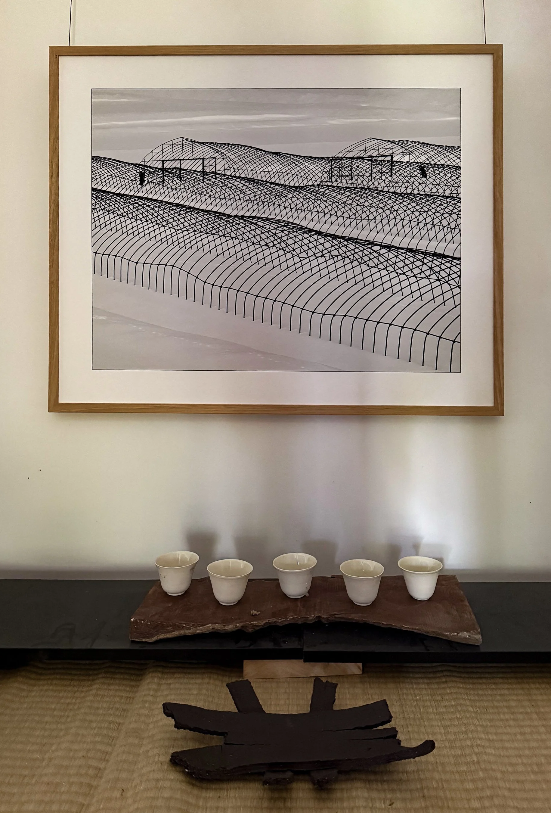 A framed black-and-white geometric artwork hangs on a white wall. Below it, a wooden plint holds five white porcela cups on a rustic wooden tray. The benc has tatami mat flooring and a dark, stoneware sculpture  in front of the shelf.