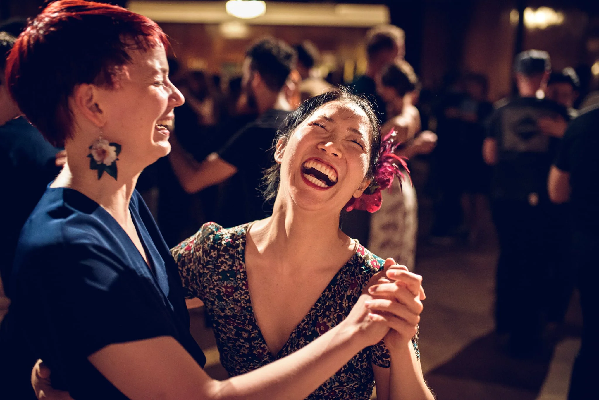 Two women laughing and dancing together at a social gathering or party, with other people dancing and socializing in the background.