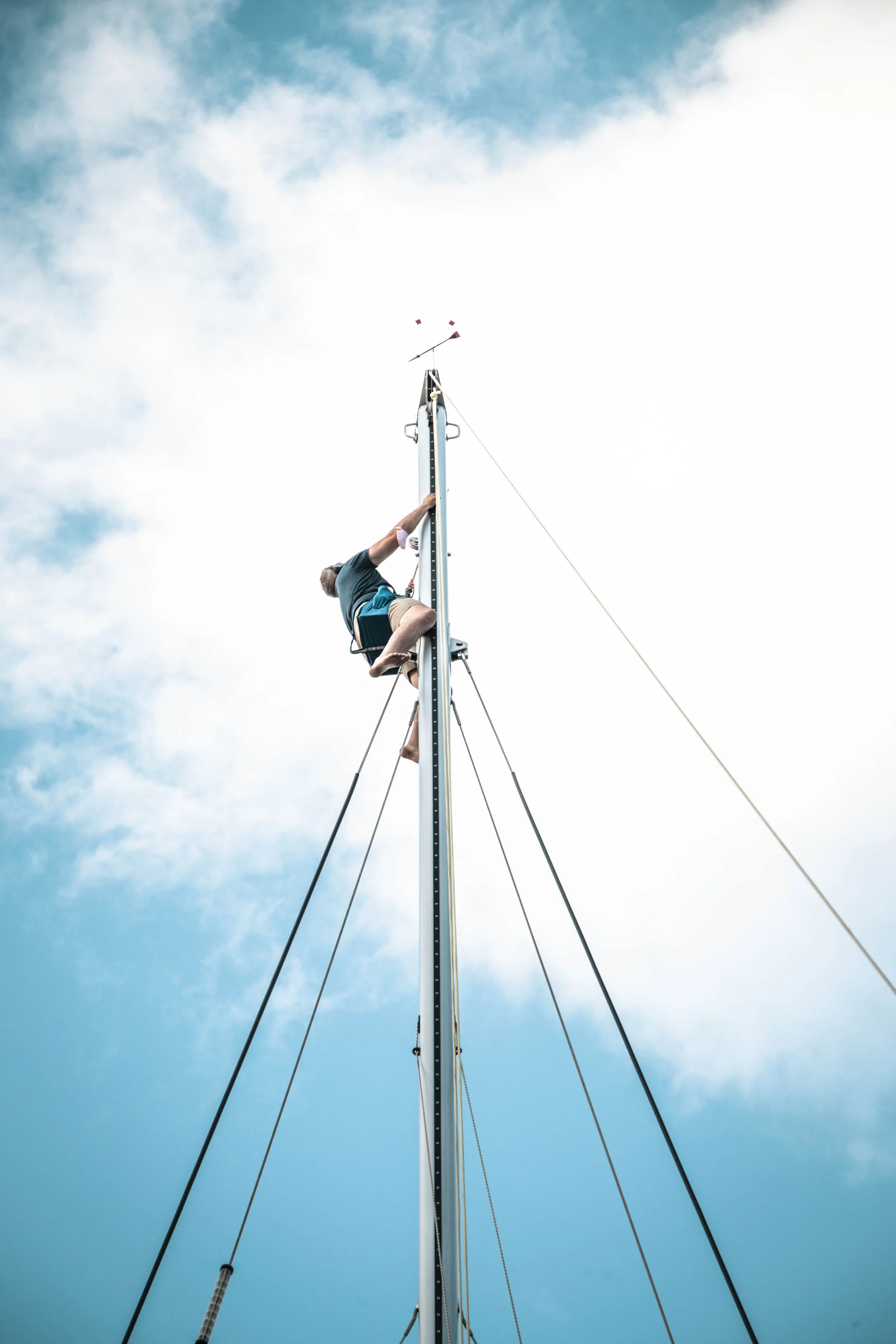 Person climbing a sailboat mast with ropes extending against a cloudy sky.