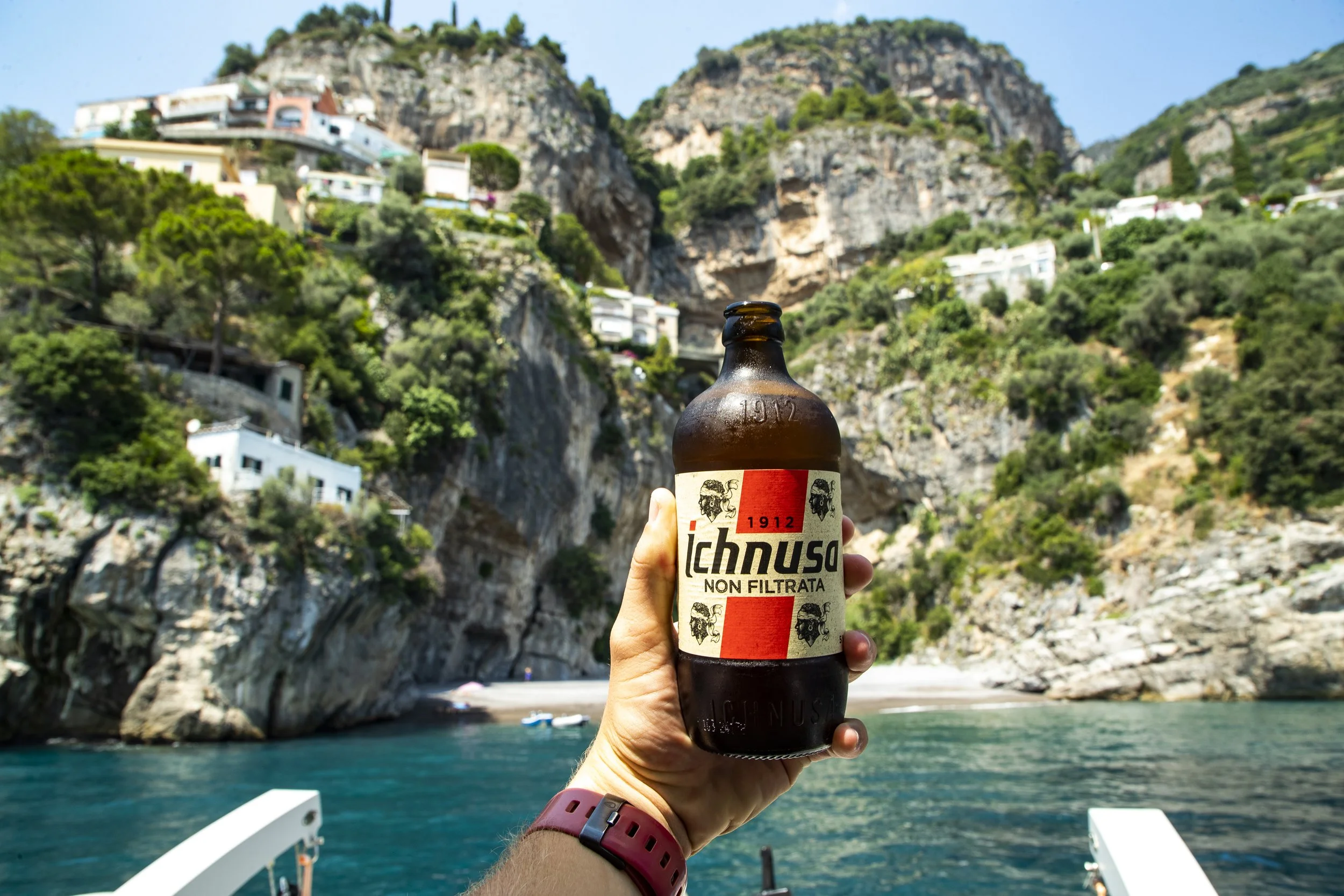 A hand holding a bottle of Ichnusa beer with a scenic coastal background of cliffs and houses.