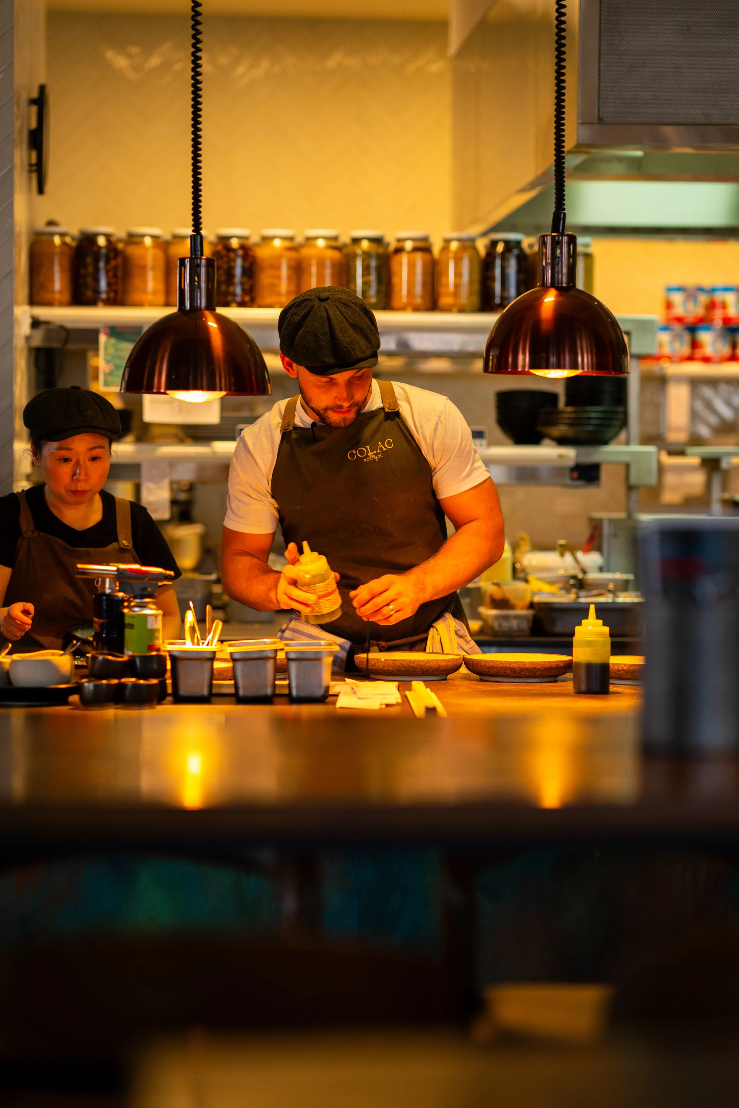 Two restaurant workers, a woman and a man, wearing black caps and aprons, are preparing food behind a counter with various condiments and kitchen tools, under copper pendant lights, in a cozy restaurant setting.
