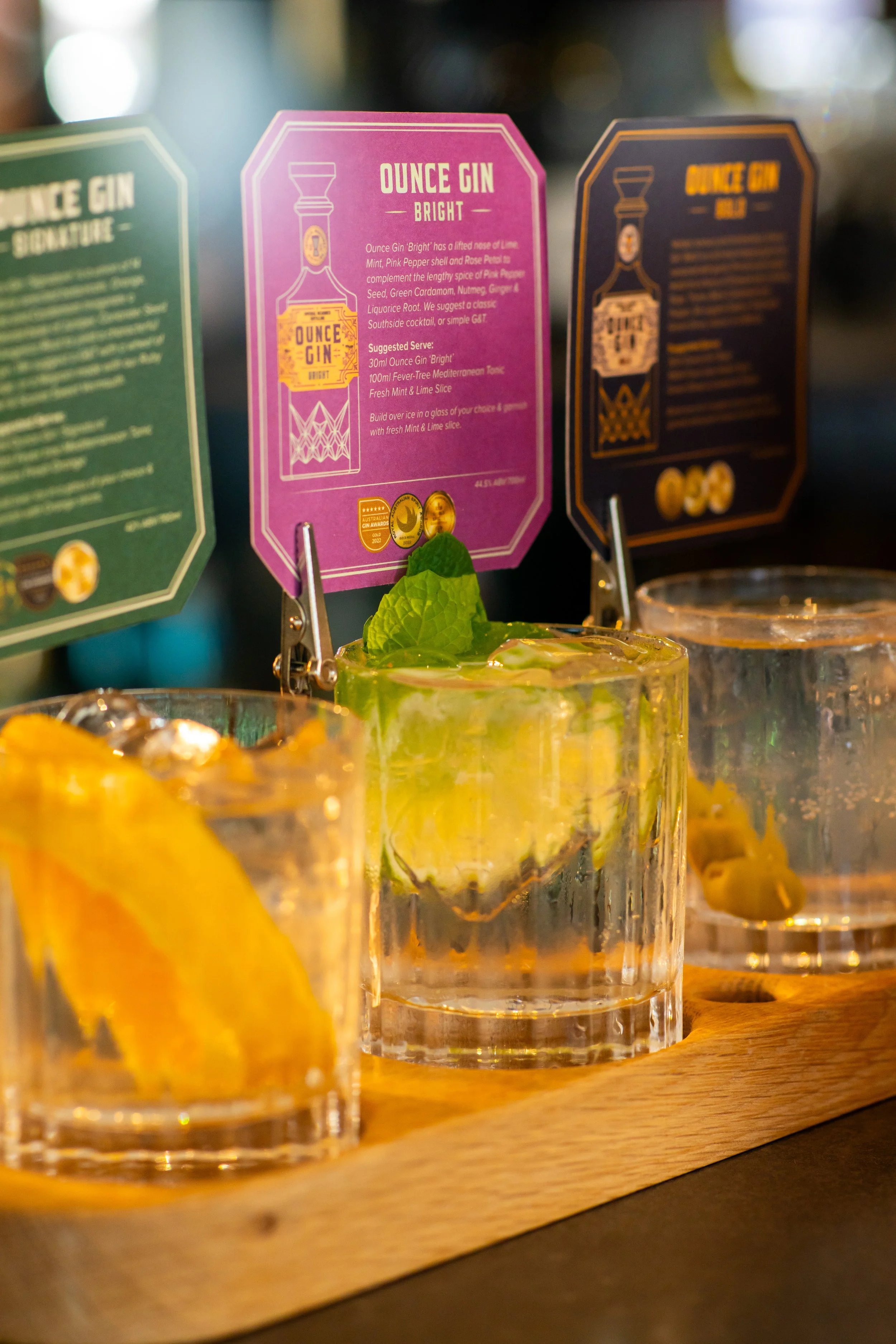 Close-up of three cocktails on a wooden tray, with colorful cocktail cards in the background, including a pink one labeled 'Ounce Gin Bright', and some mint leaves and orange slices visible in the drinks.