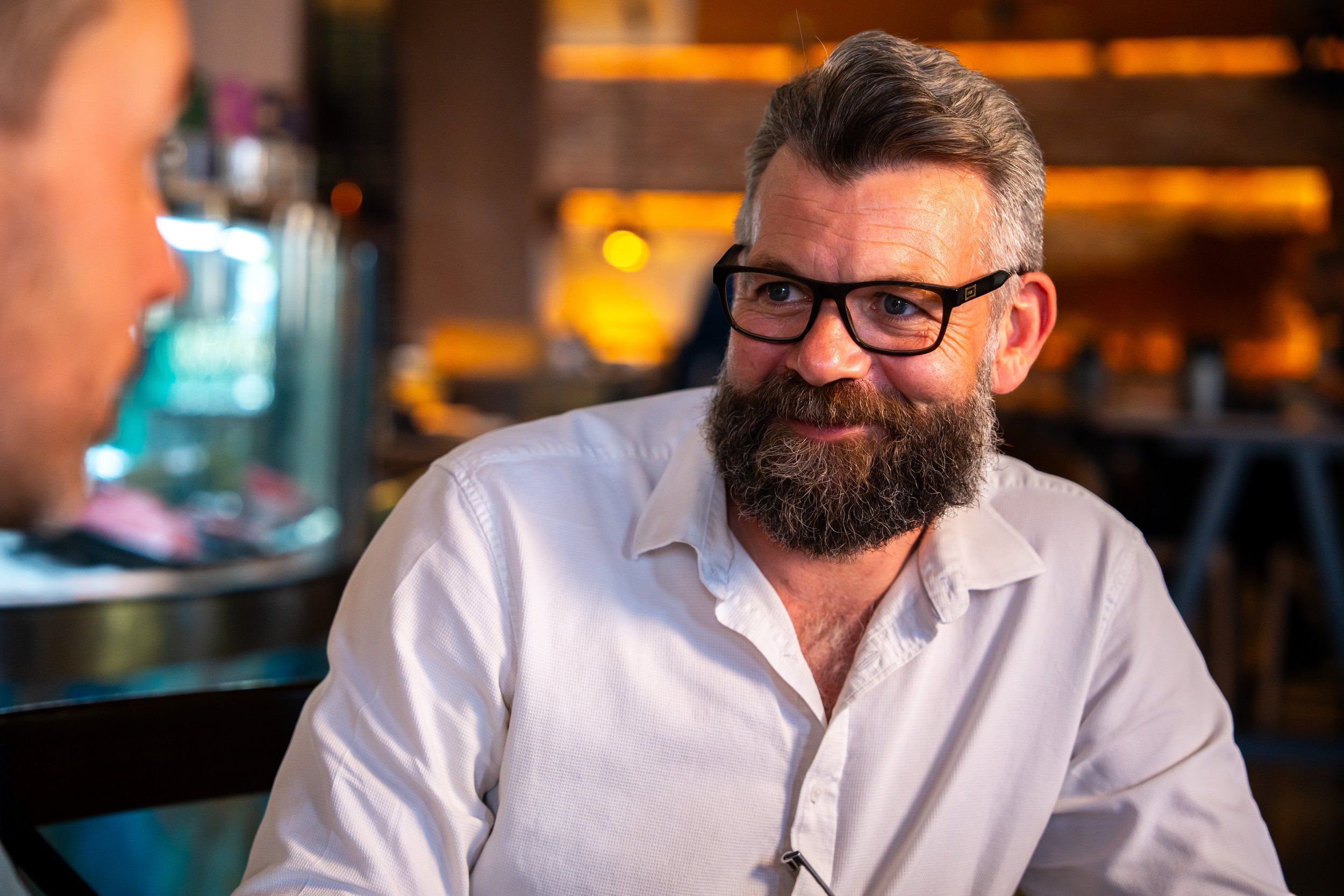 Man with gray hair, beard, and glasses smiling and talking to someone in a casual setting with warm lighting.