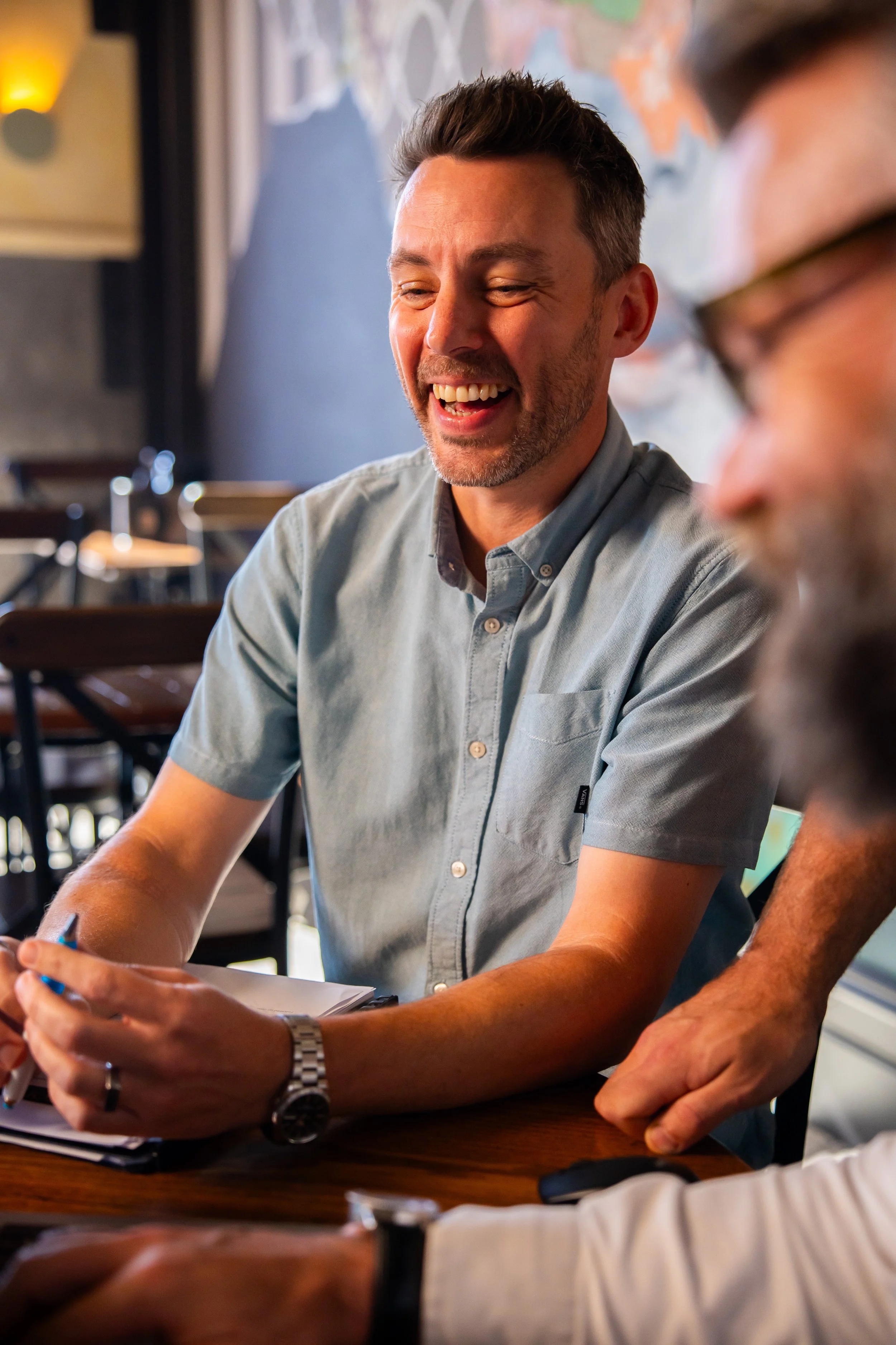 Two men sitting at a table engaged in conversation, smiling and laughing in a brightly lit indoor setting.