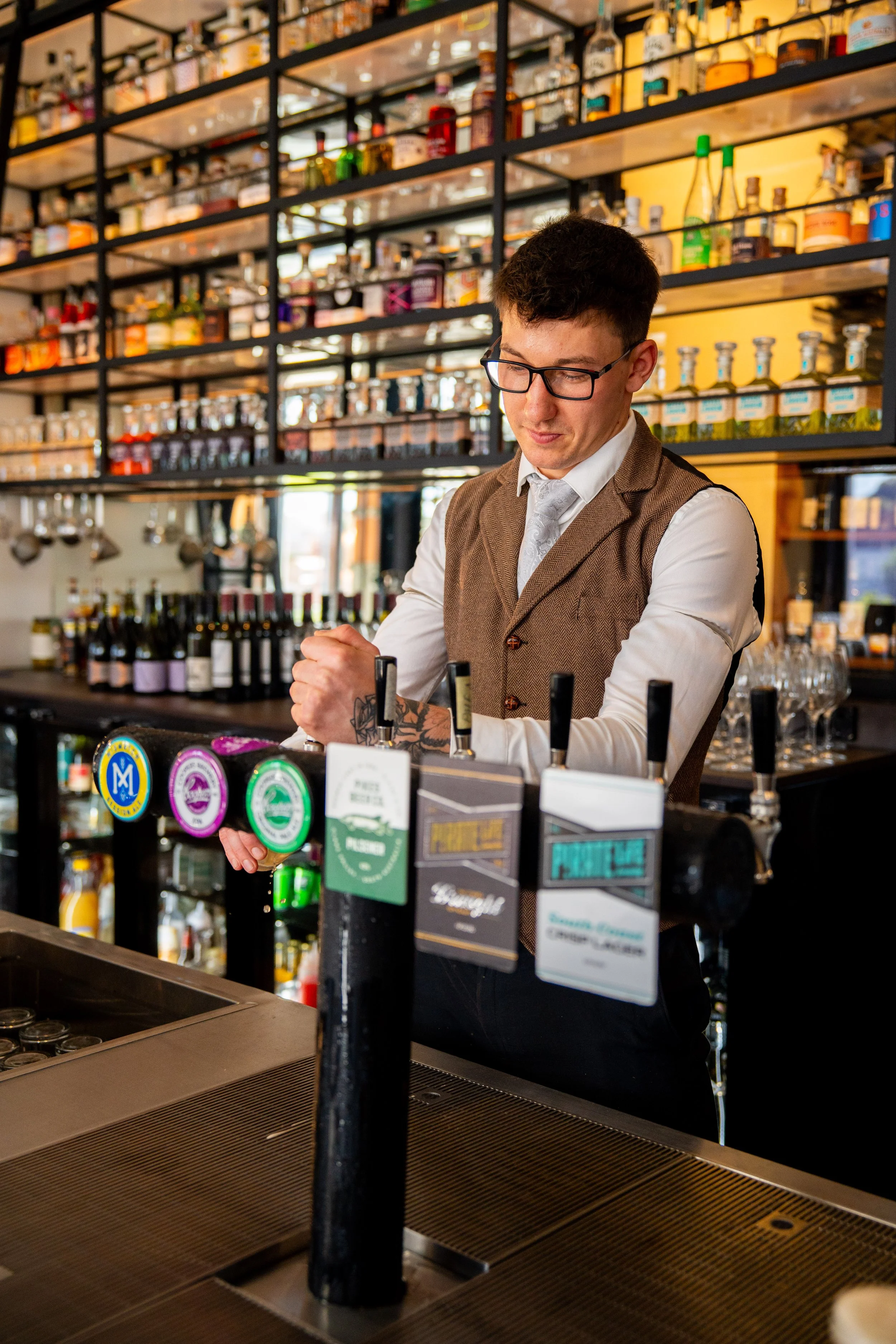 A bartender wearing glasses and a brown vest is working behind the bar, surrounded by bottles and beer taps in a bar or pub.