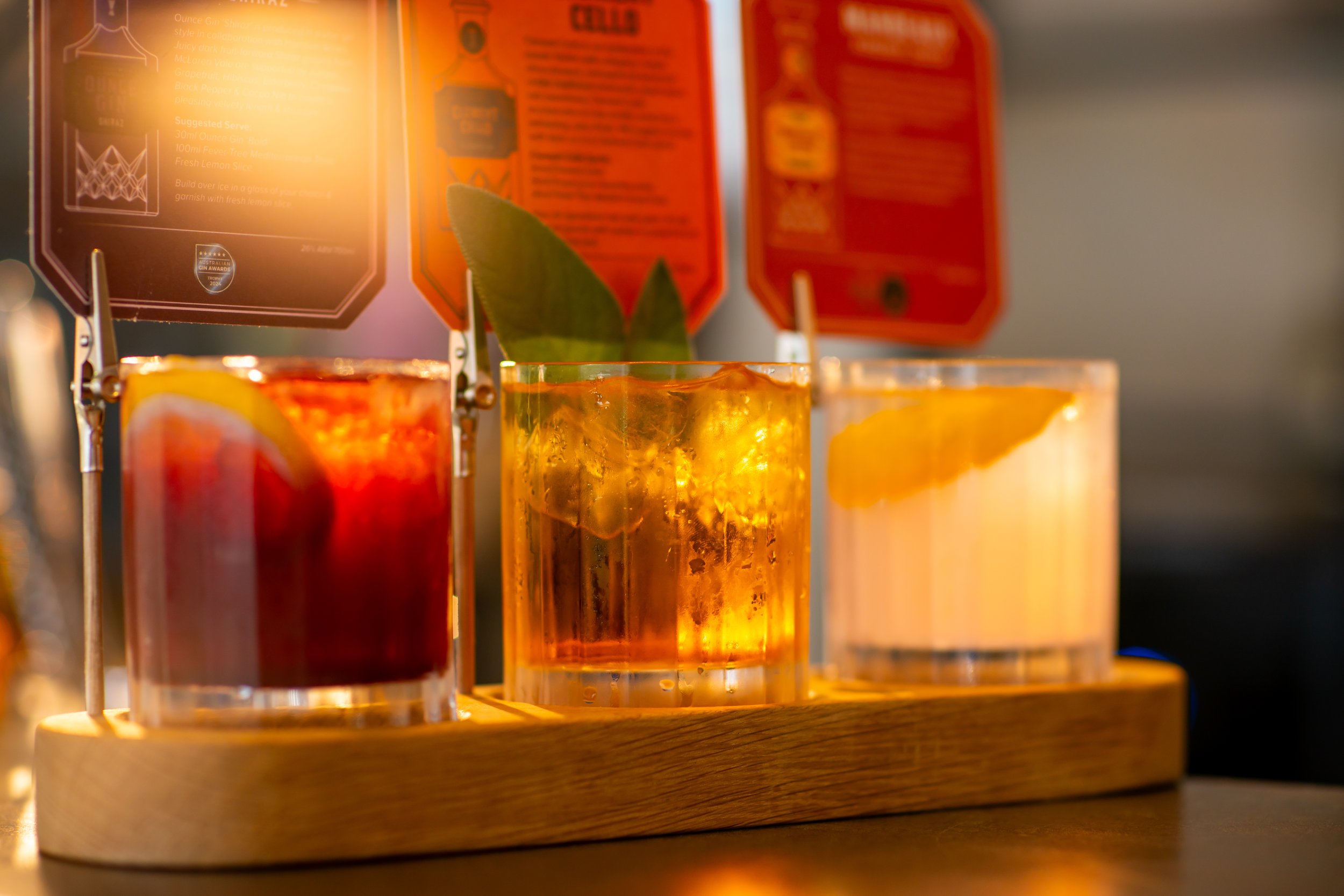Three tall glasses filled with colorful beverages and ice, set on a wooden tray, with orange and red packaging in the background.