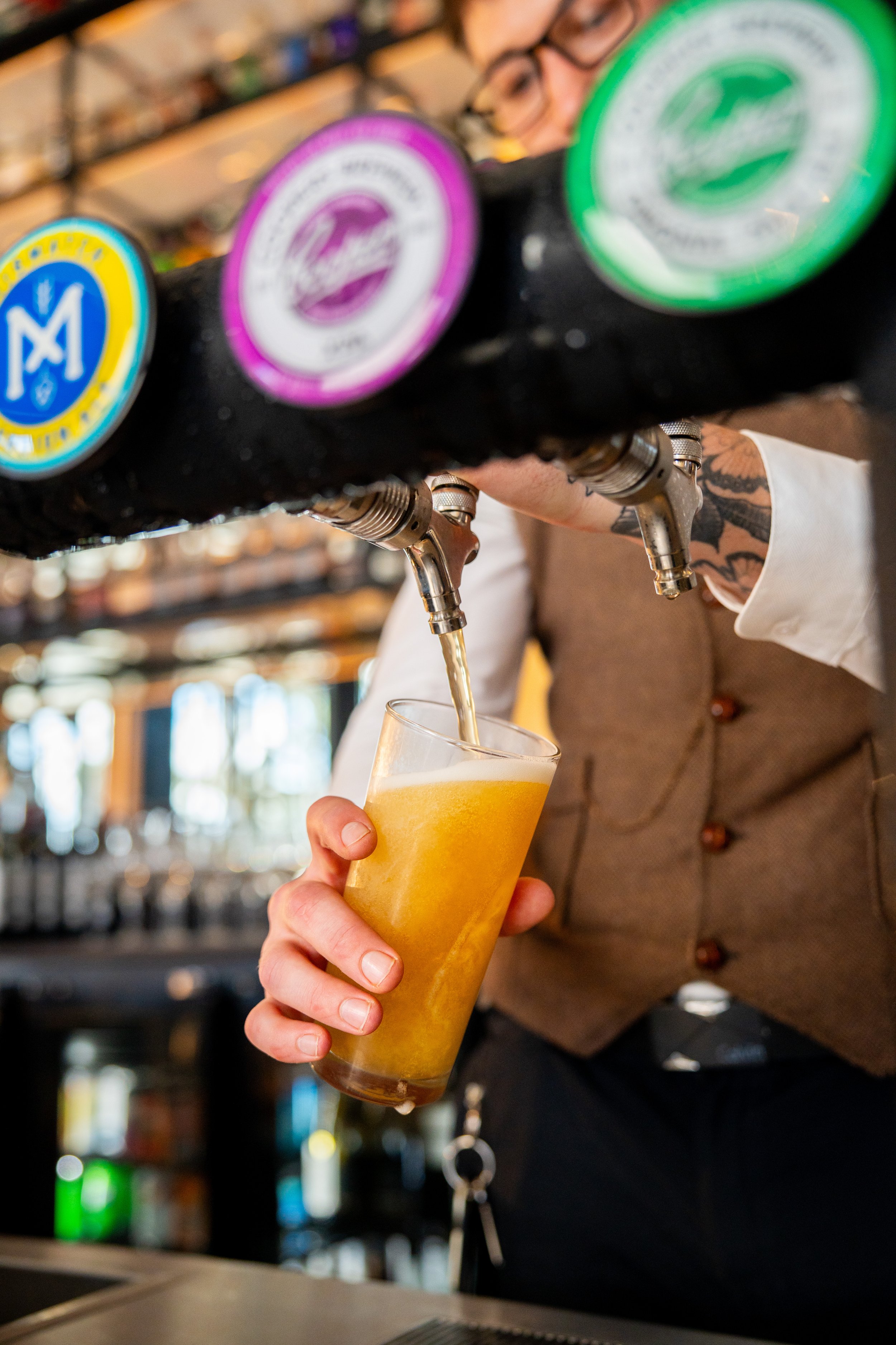 A bartender is pouring a draft beer from a tap into a glass, with colorful tap handles and blurred shelves of bottles in the background.