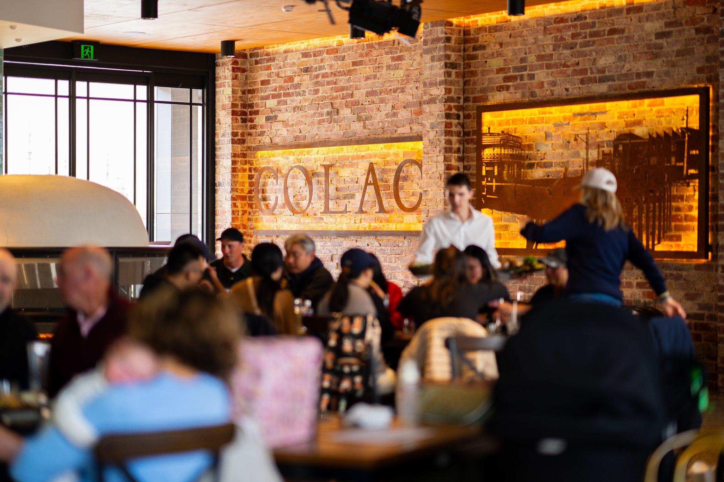 A restaurant with a rustic brick wall and large windows. The wall displays illuminated text reading "COLAC". People are dining, and waitstaff are serving in a lively atmosphere.