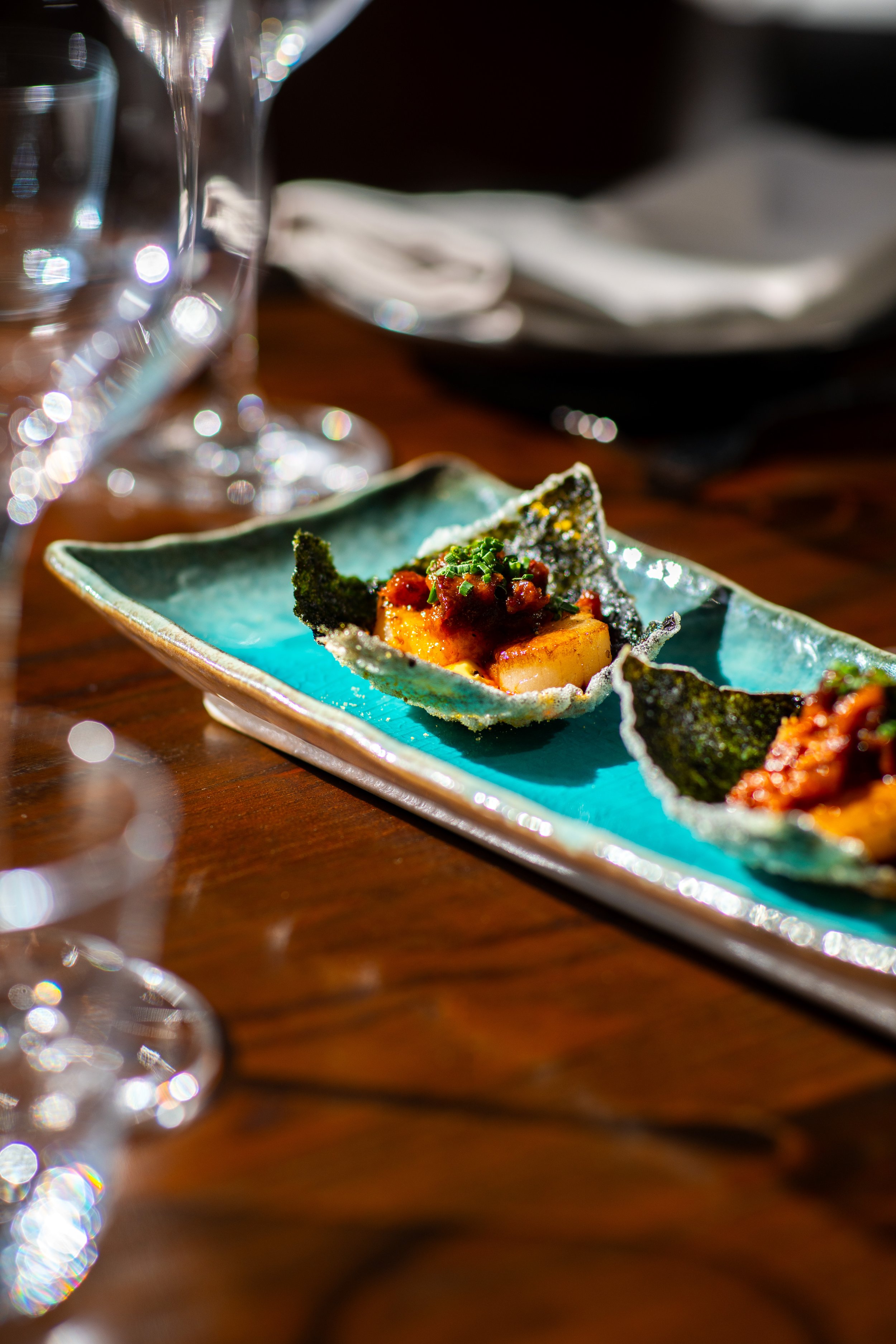 Close-up of a small, rectangular plate with a piece of fish topped with sauce and chopped herbs. In the background, blurred glasses and a dark table setting.