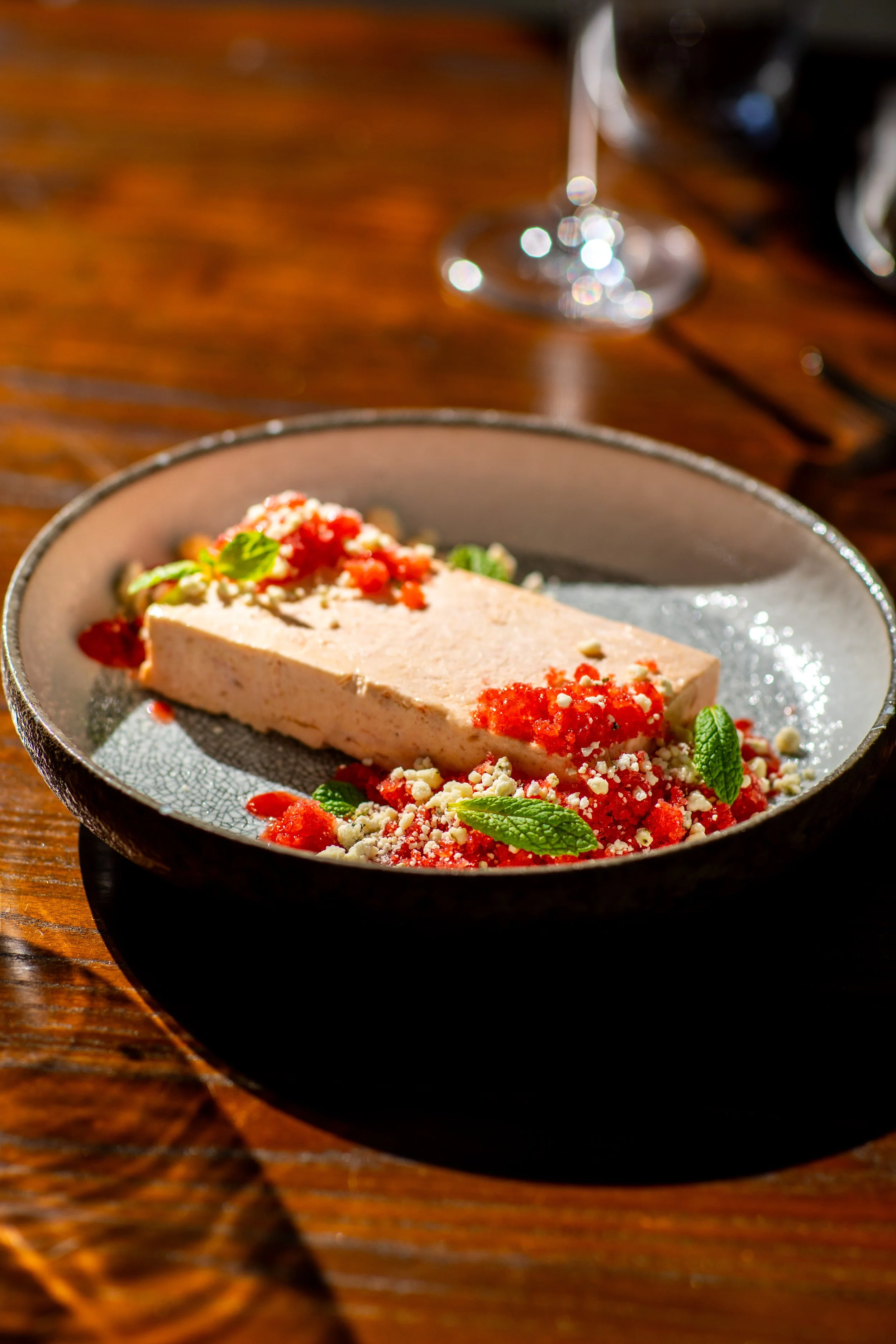 A slice of pie on a plate with red fruit topping, garnished with mint leaves, inside a black bowl on a wooden table, with a blurred wine glass in the background.