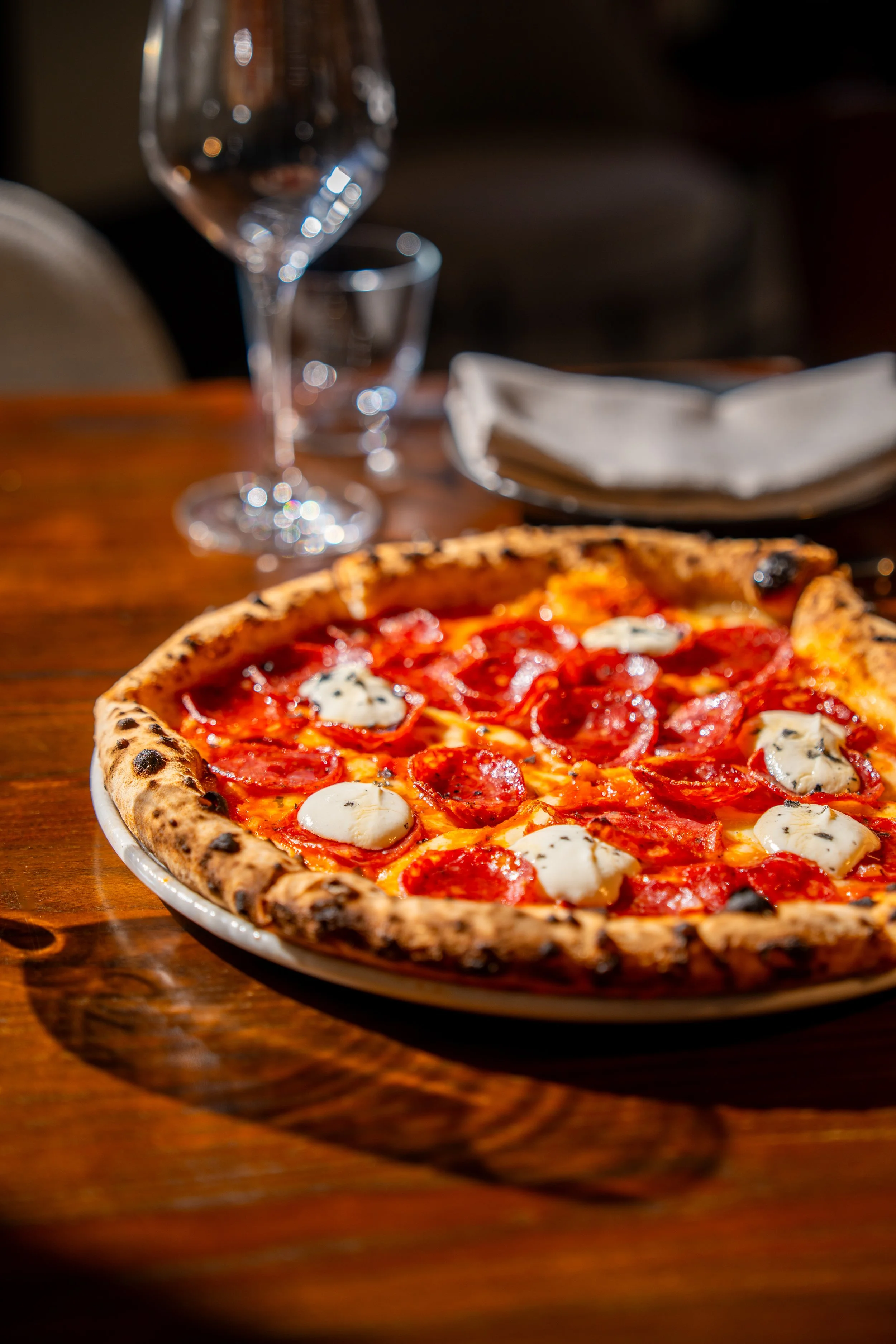 Close-up of a pepperoni and blue cheese pizza on a wooden table with an empty wine glass and a napkin in the background.