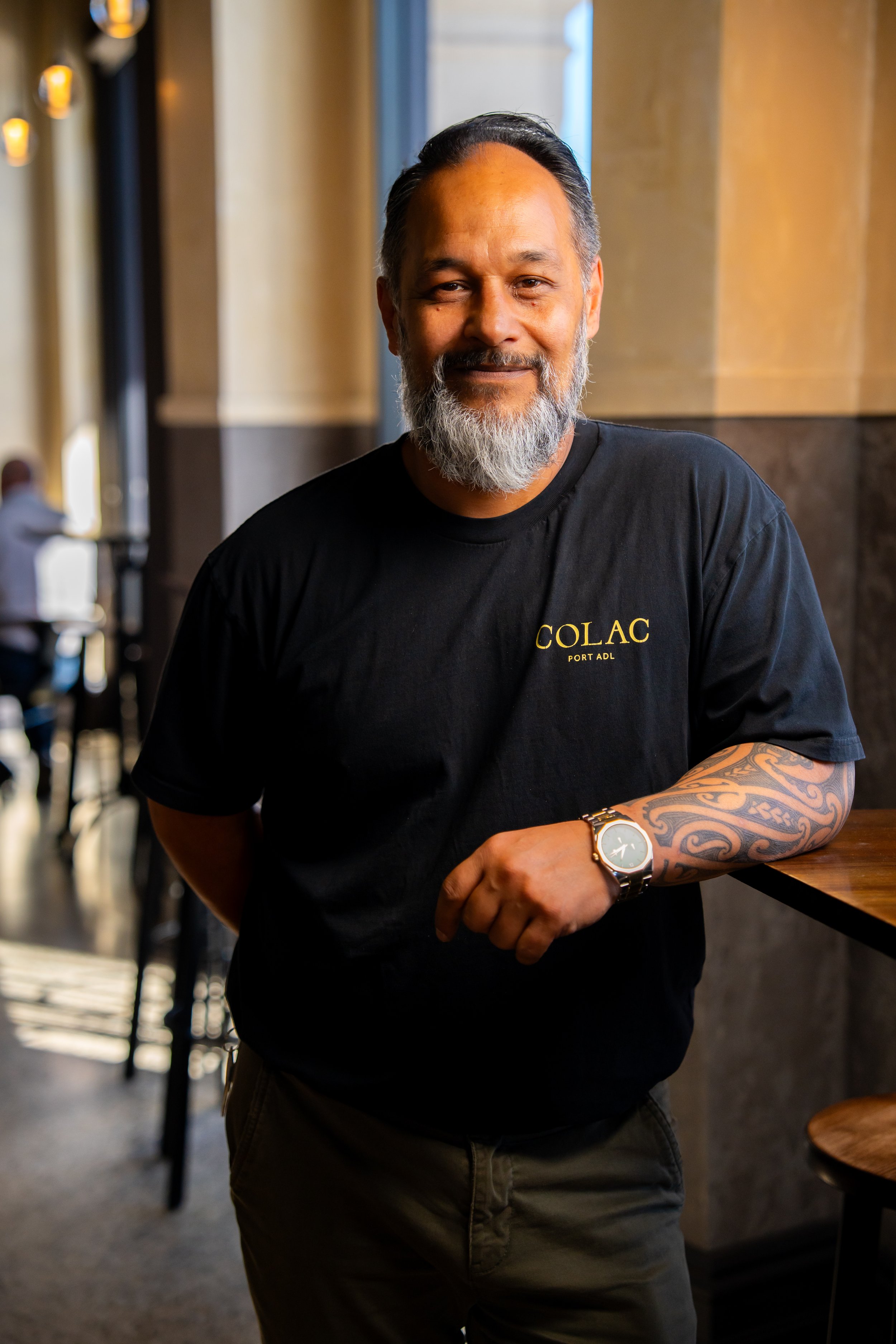 A middle-aged man with a beard, wearing a black T-shirt with 'COLAC PORT ADL' on it, standing inside a cafe or restaurant, smiling, with a tattooed arm and a wristwatch.