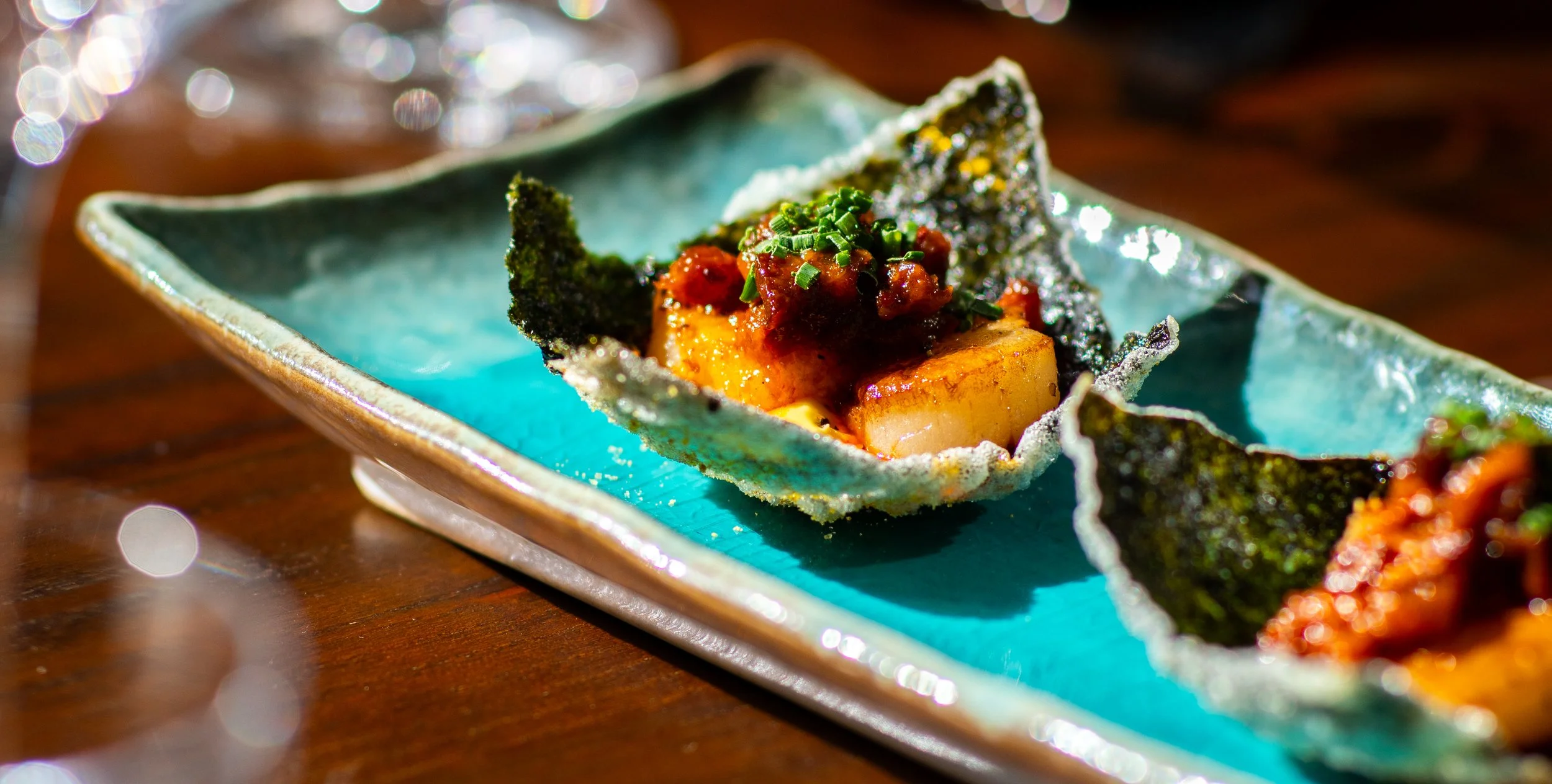 Close-up of a gourmet appetizer featuring a seared scallop topped with a spicy tomato-based sauce and chopped green herbs, served on a decorative ceramic dish.