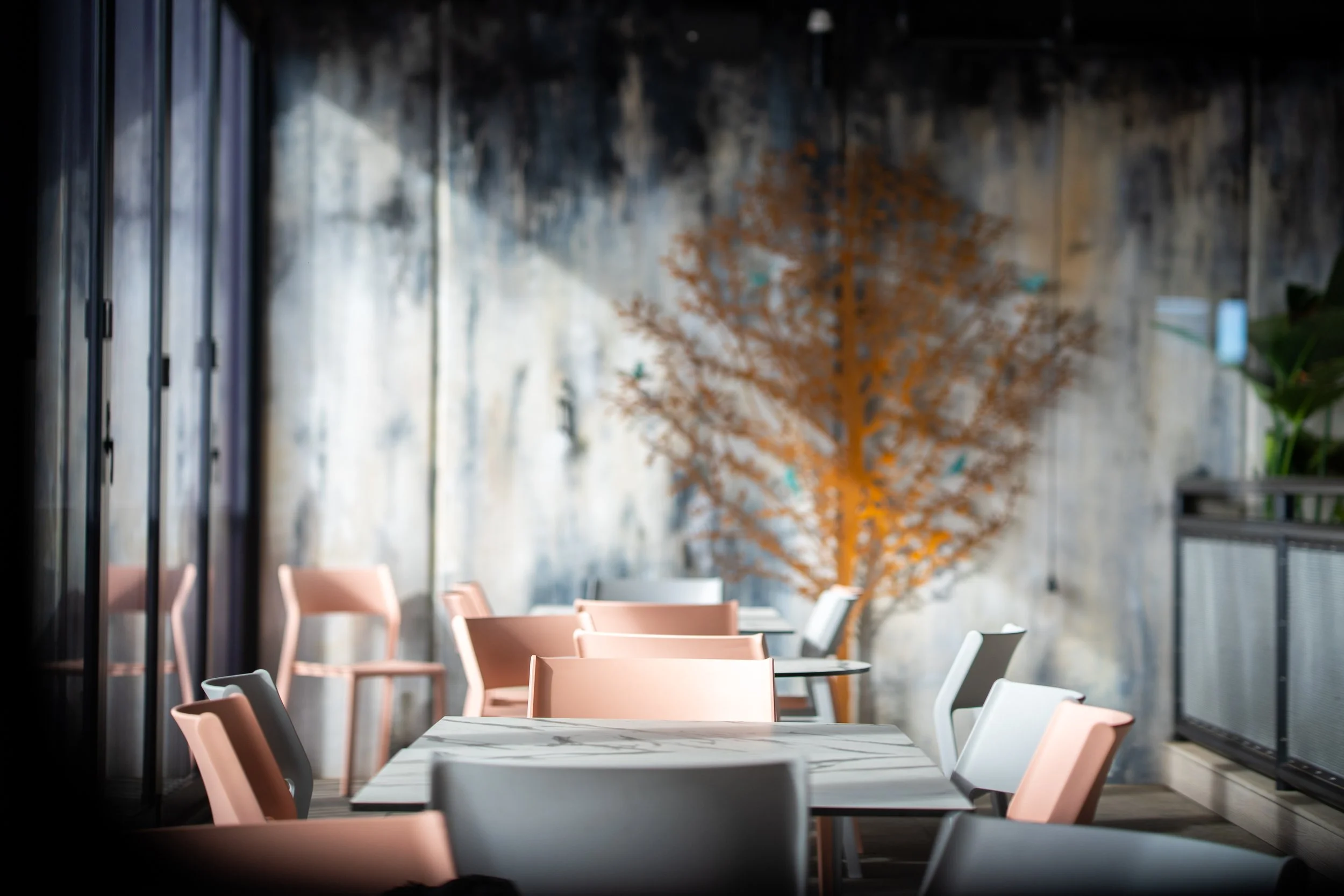 Interior of a modern cafe or restaurant with pink and white chairs around a marble table, a large window on the left, and a decorative orange tree in the background.