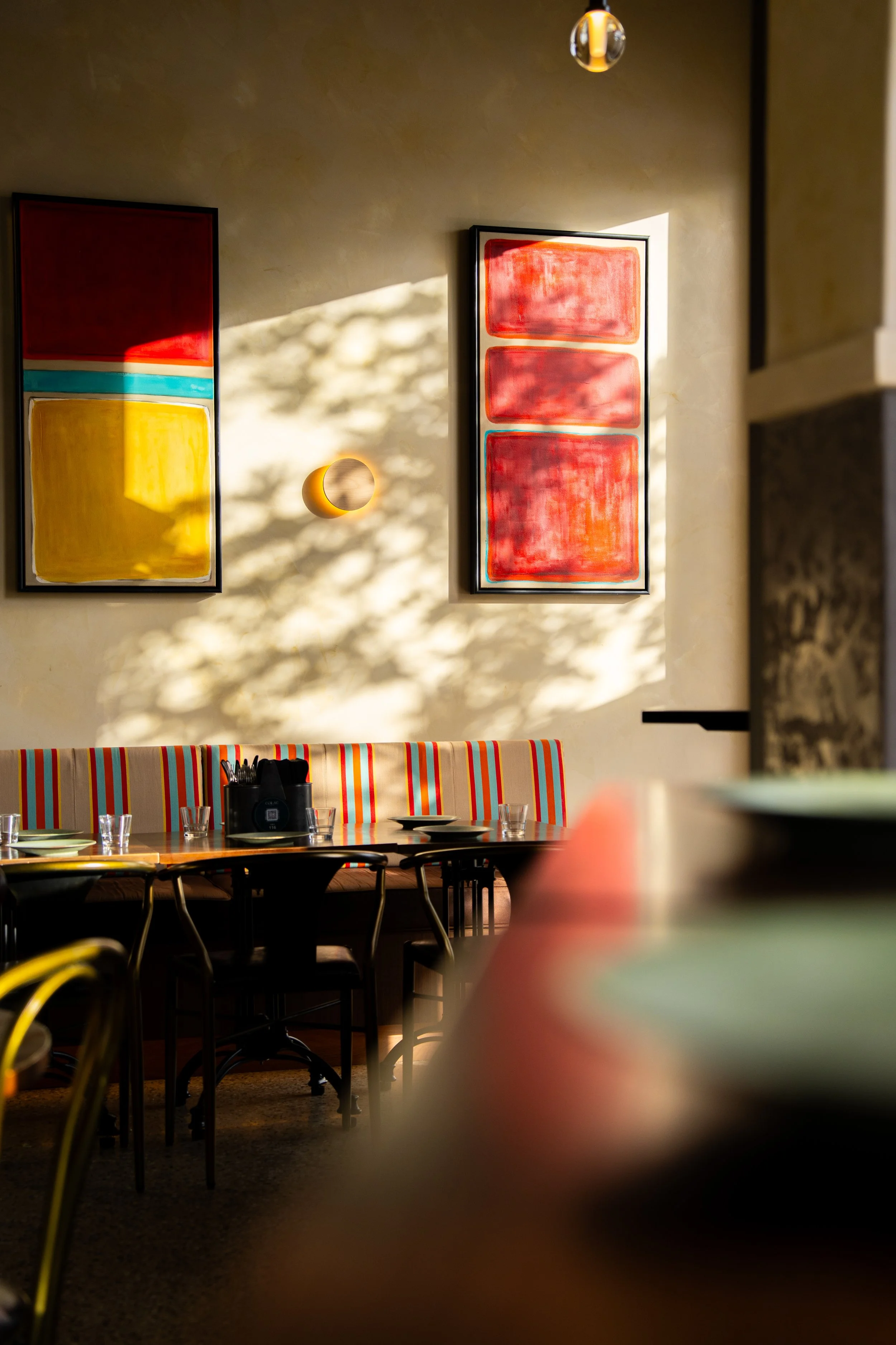 An indoor dining area with a wooden table set with glasses and utensils. There is a colorful, striped bench and chairs around the table. Behind, two abstract paintings with red, yellow, and turquoise colors are hung on a beige wall, with sunlight cas
