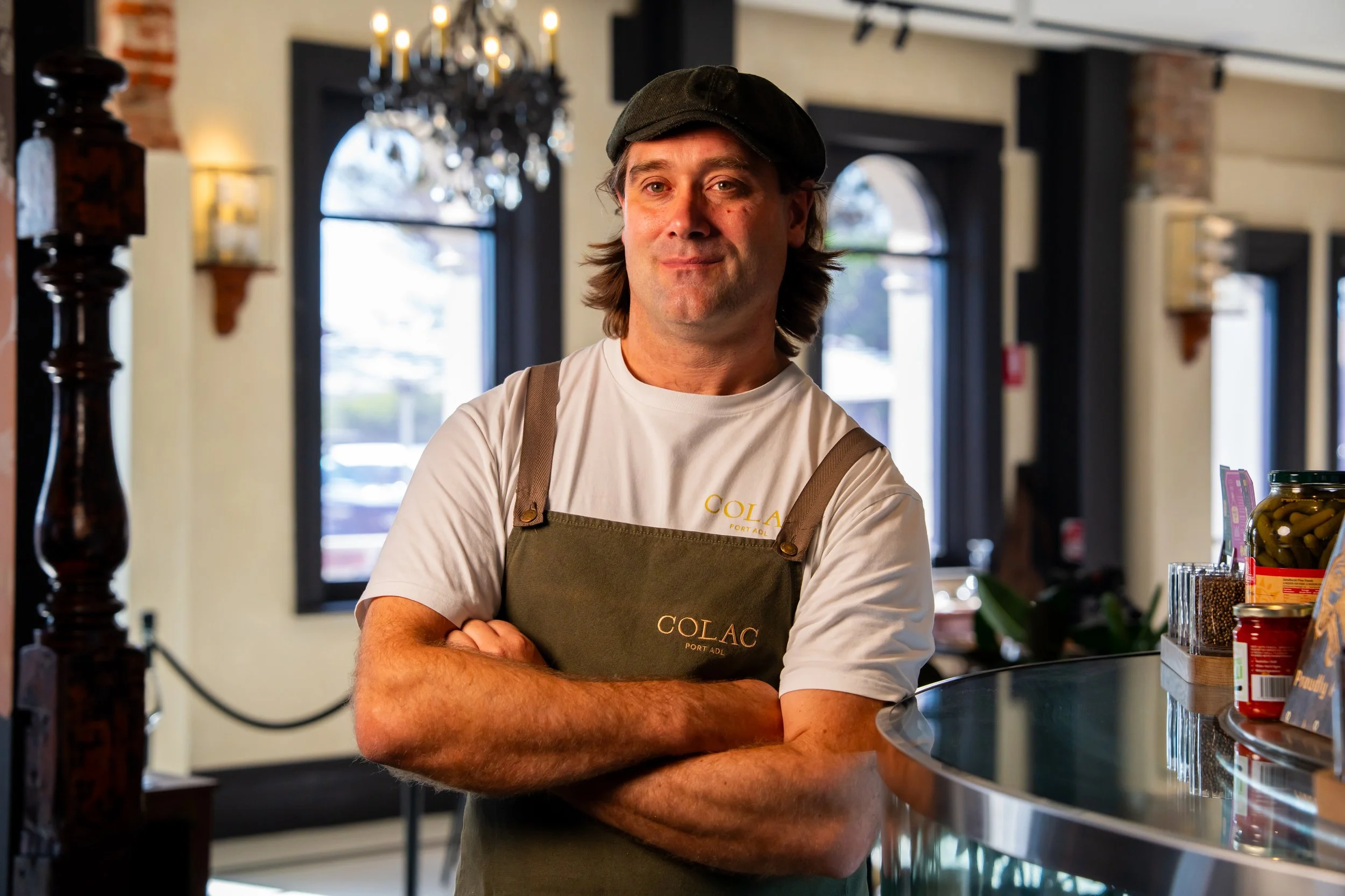 A man wearing a black cap and a gray apron with the word 'GOLAC' printed on it, standing with his arms crossed inside a store or cafe with large windows, a chandelier, and shelves with products behind him.