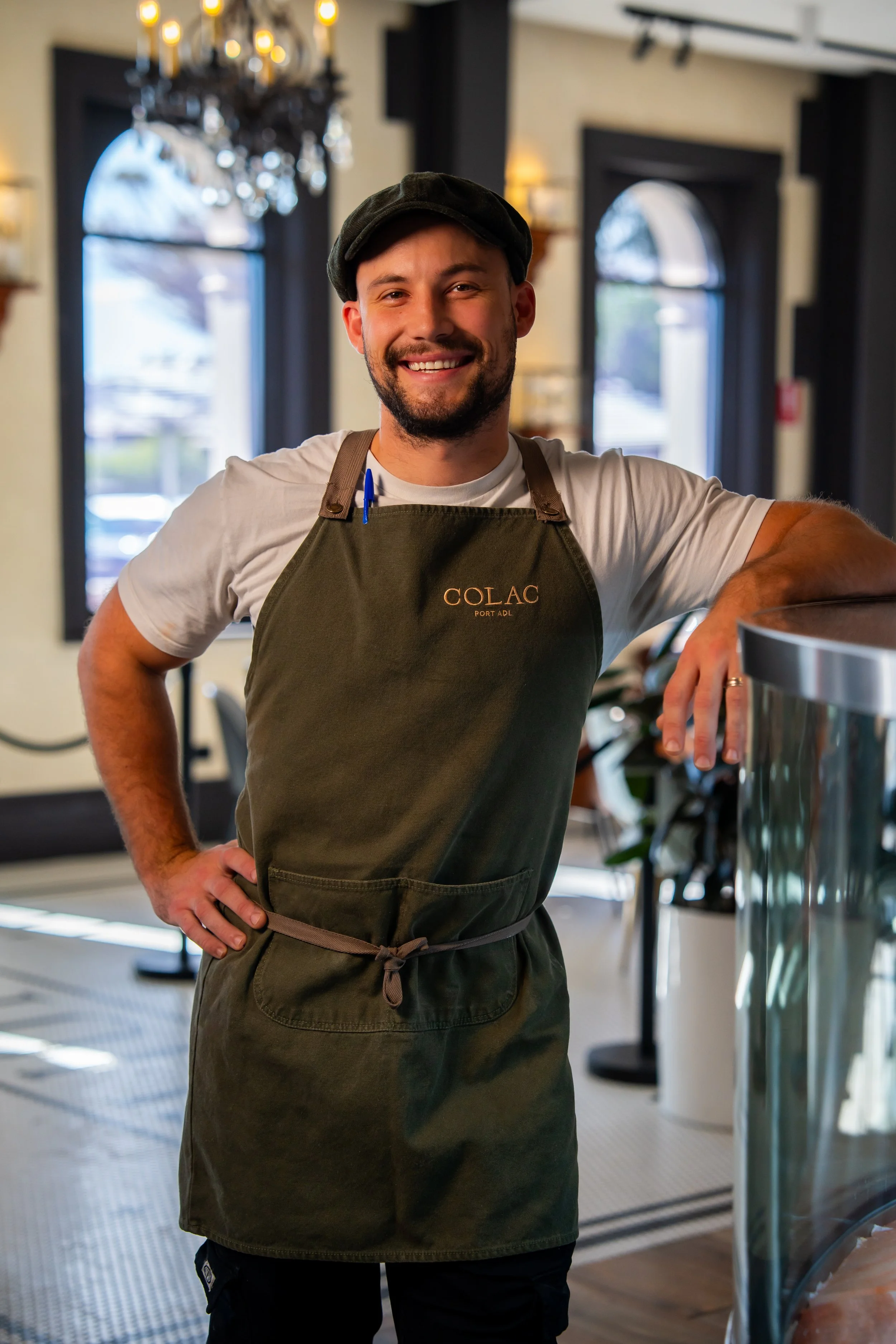 Smiling man in a restaurant or café, wearing a black cap, white t-shirt, and green apron with the word 'COLAC' on it. He is standing with one hand on his hip and the other resting on a counter.