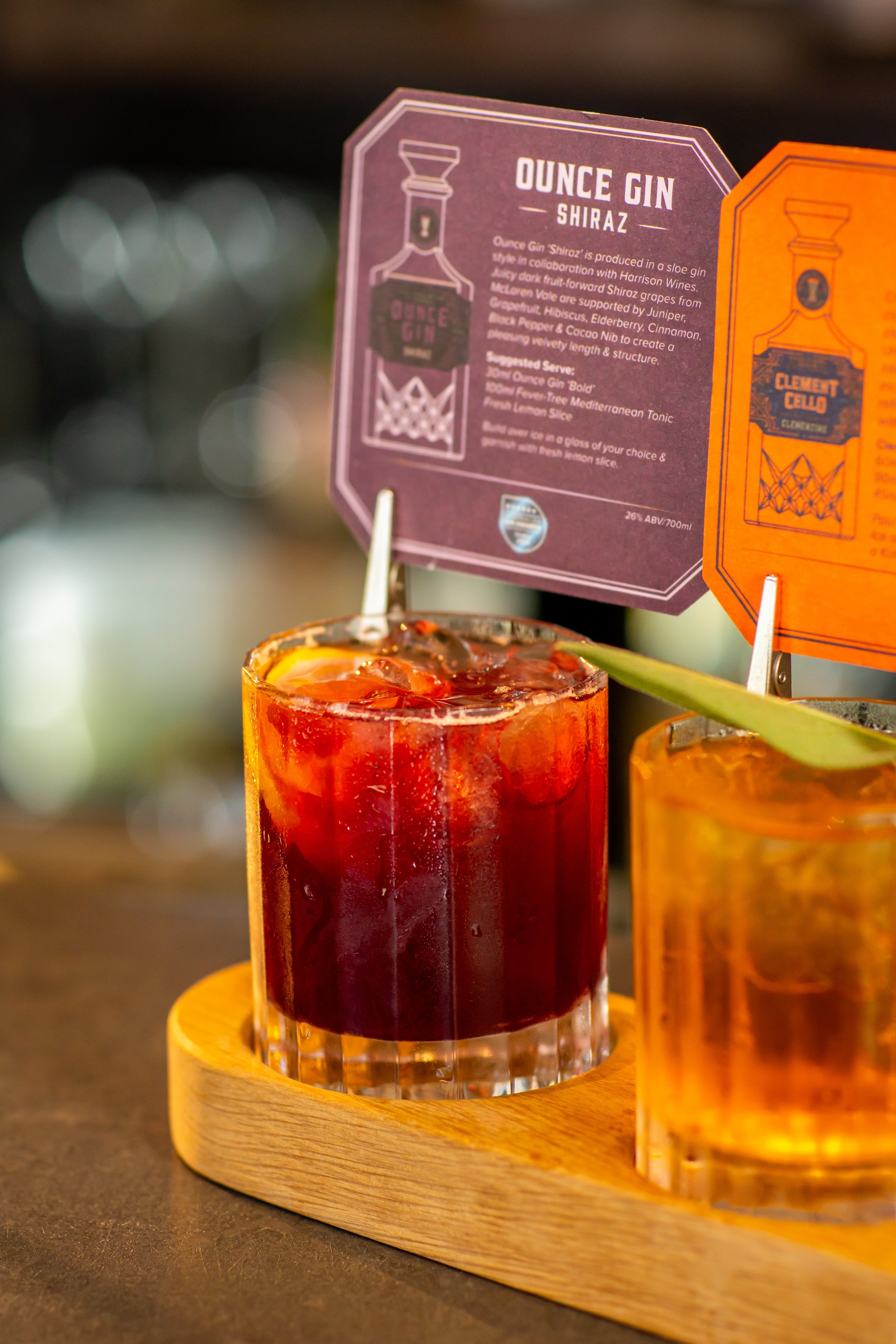 A close-up of a menu card for a cocktail called 'Ounce Gin' Shiraz, positioned behind a glass filled with a dark red beverage on a wooden serving tray. The tray is placed on a dark surface, with blurry bokeh lights in the background.