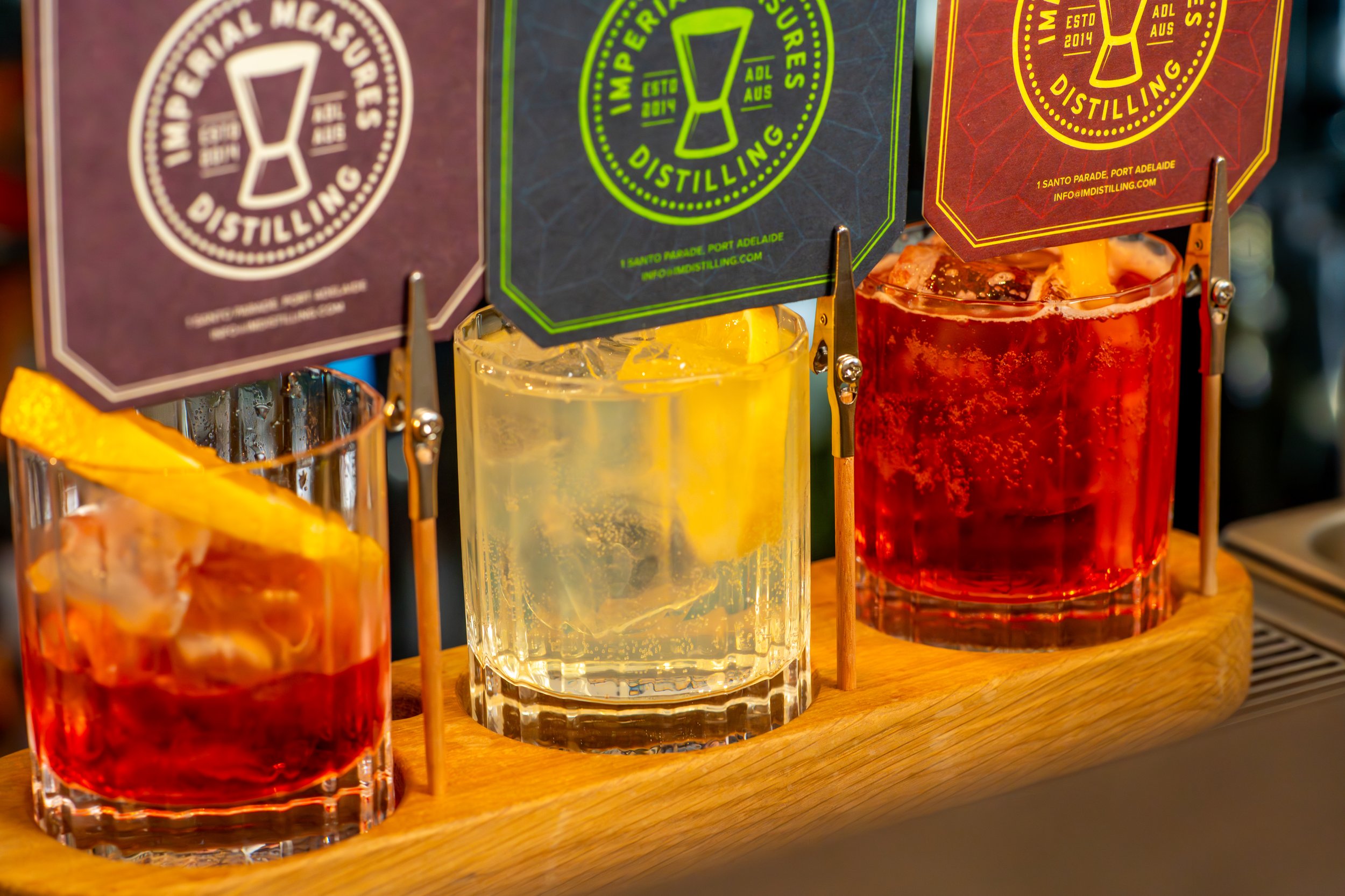 Three glasses with colorful drinks lined up on a wooden surface, with colorful packaging in the background. The drinks are red, yellow, and a clear liquid with ice.