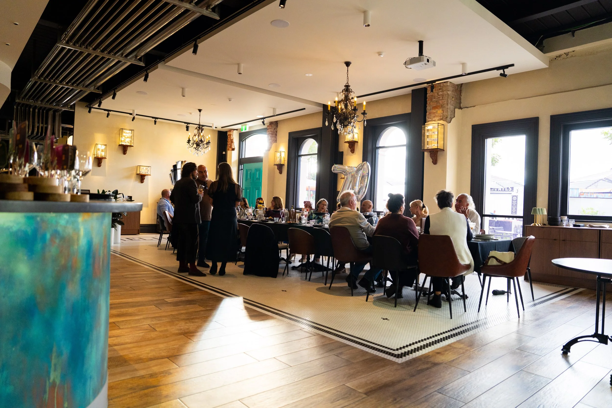 The image shows a restaurant interior with a group of people dining and socializing. There are large windows, chandeliers, and decorative wall sconces providing warm lighting. In the foreground, part of a colorful table or counter is visible.