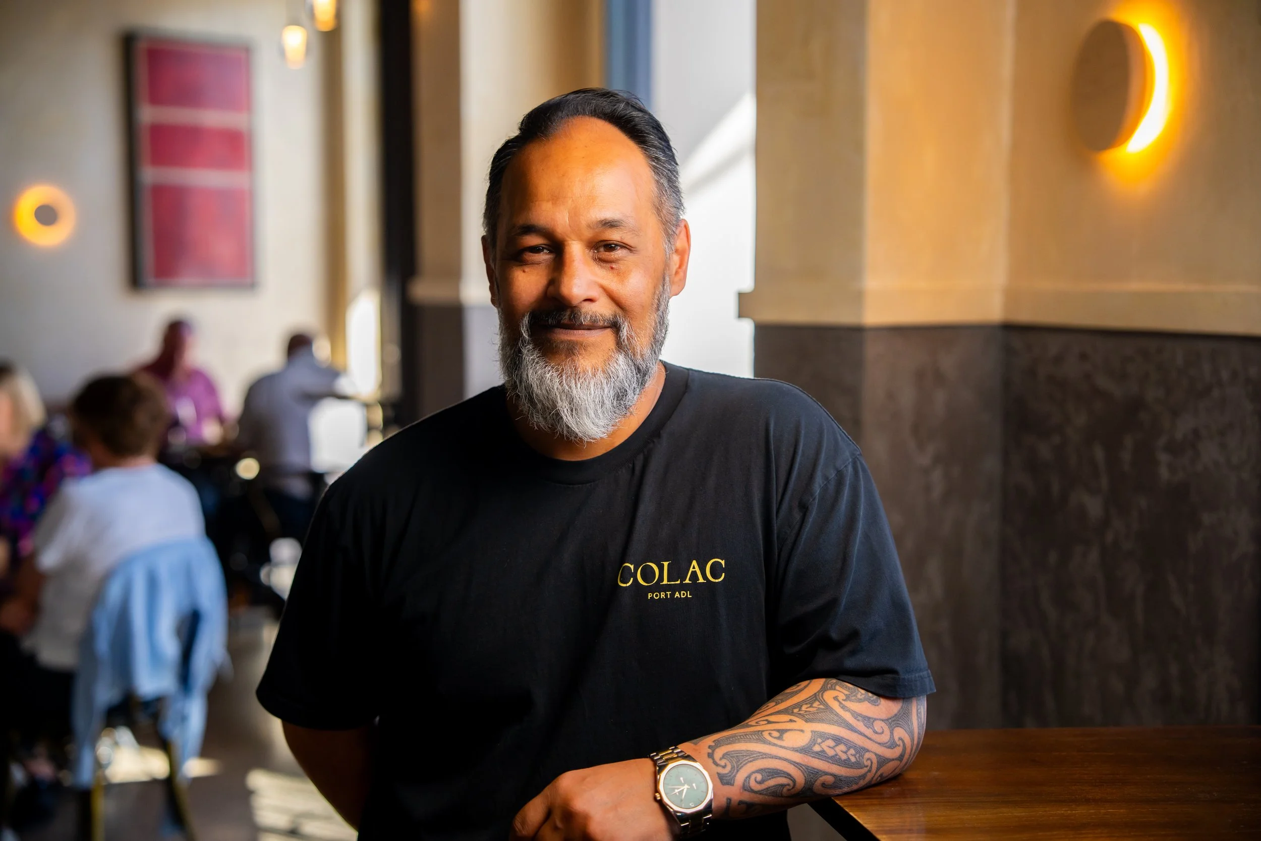 A middle-aged man with a beard and tattoos on his arm, wearing a black t-shirt and a watch, standing in a restaurant or cafe with a warm ambiance and blurred patrons in the background.