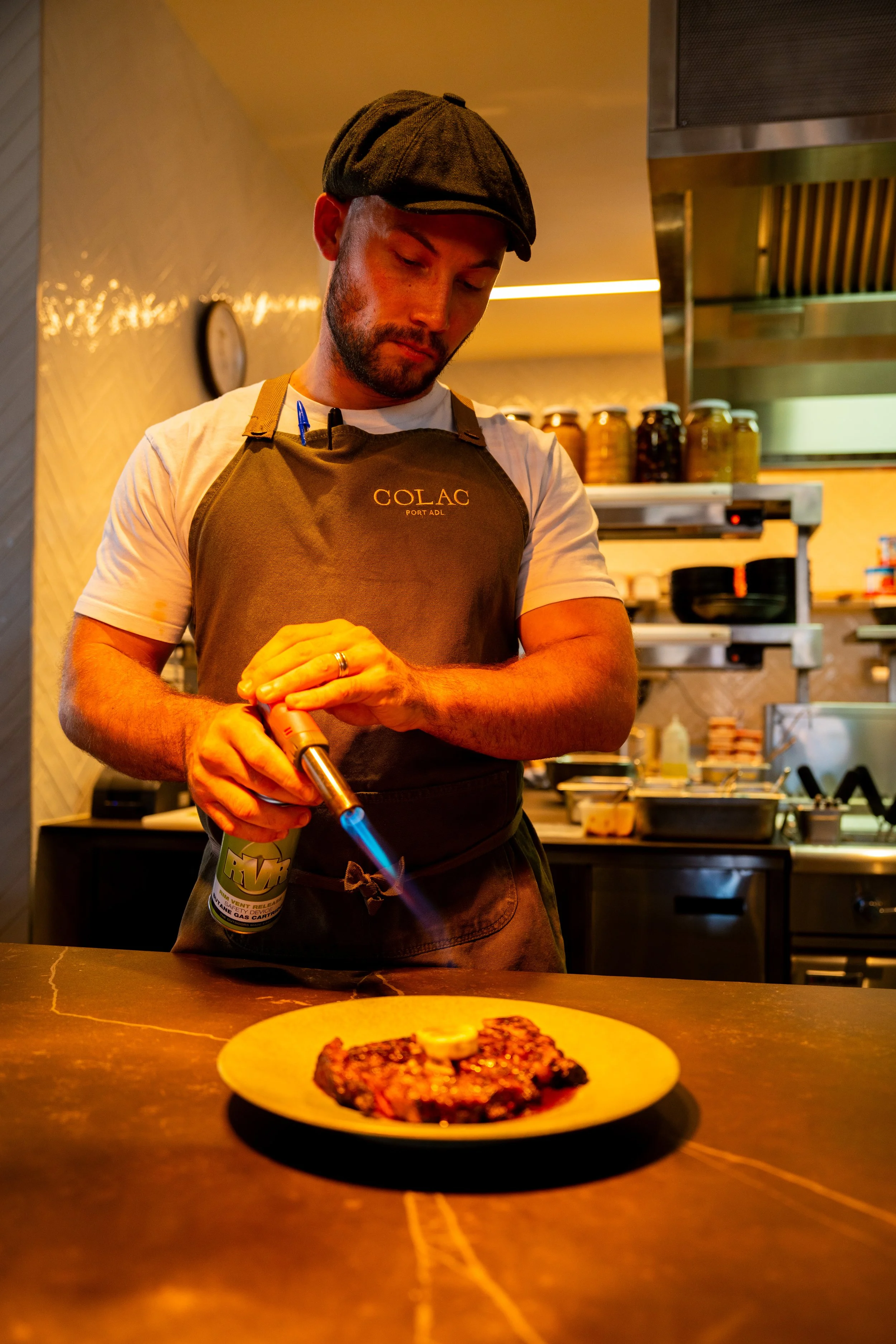 A man in a black cap and apron is working in a kitchen, holding a blowtorch and preparing to caramelize a dish on a plate with food in front of him. Behind him are jars, shelves, and kitchen equipment.