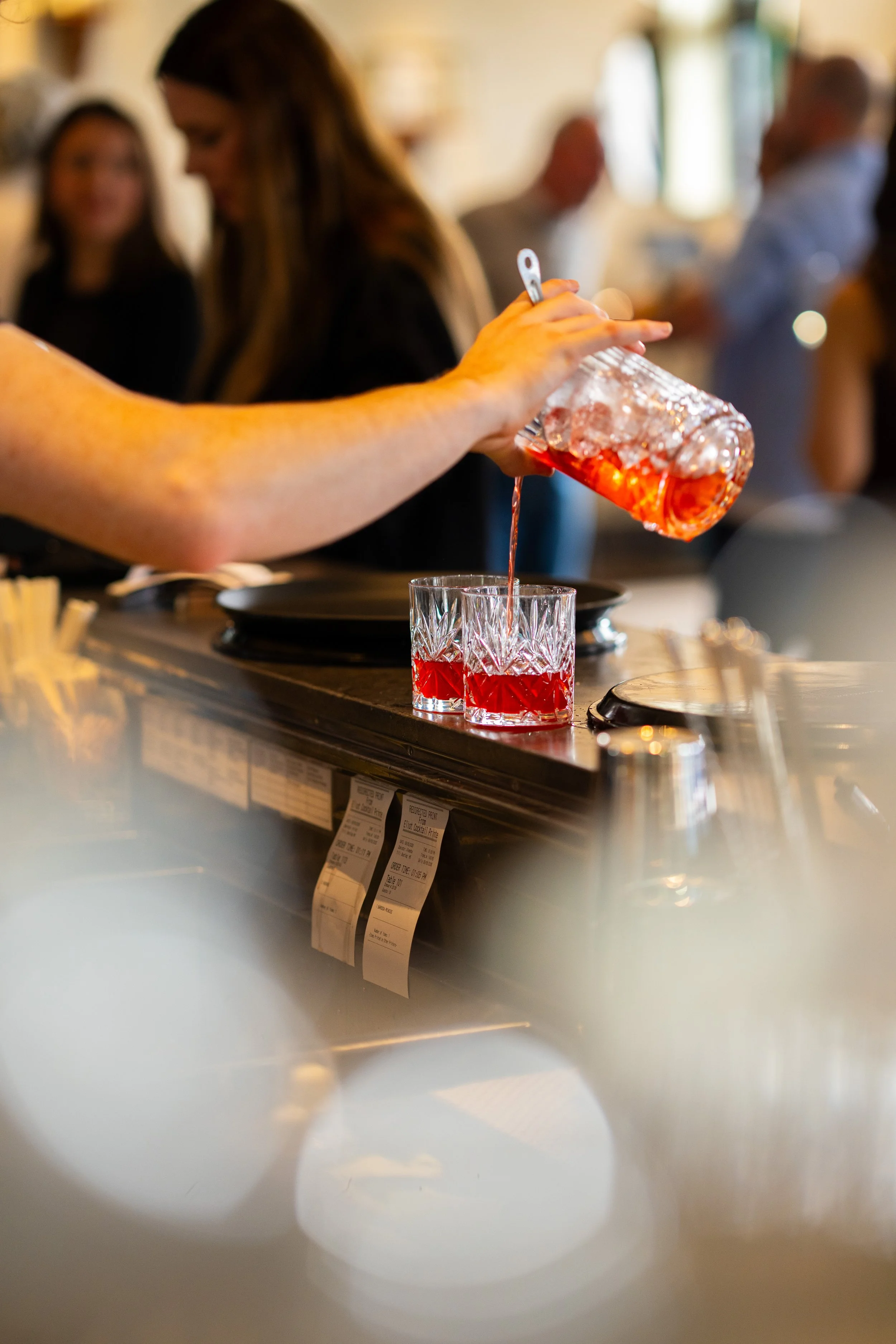 A person holding a glass of red beverage garnished with ice, with other people in the background.