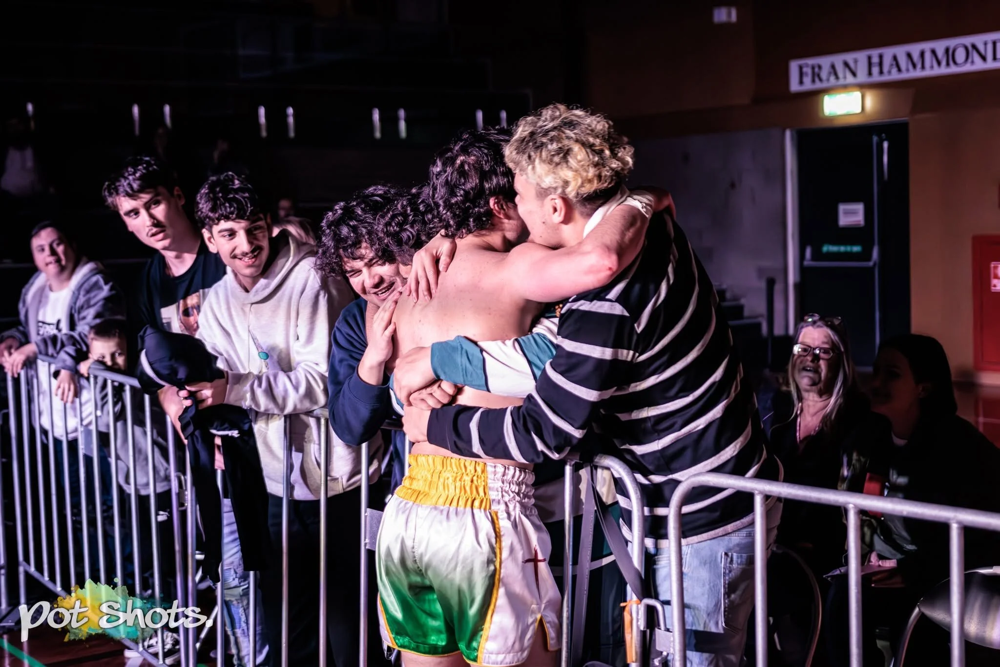 Two men embracing in front of a group of people behind a barrier at an indoor event, with one man shirtless and the other in a striped shirt. Others are watching, some smiling. Sign reads 'FRAN HAMMOND' in the background.