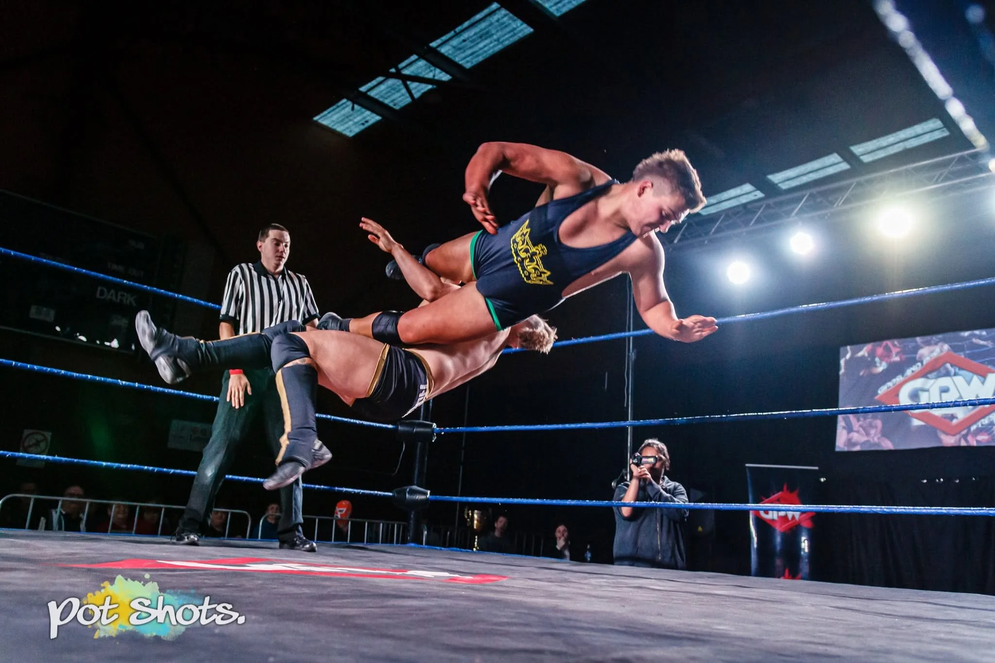 Two professional wrestlers competing in a wrestling match inside a ring, with one wrestler lifting the other in a suplex move. A referee and an audience are visible in the background under bright stadium lights.