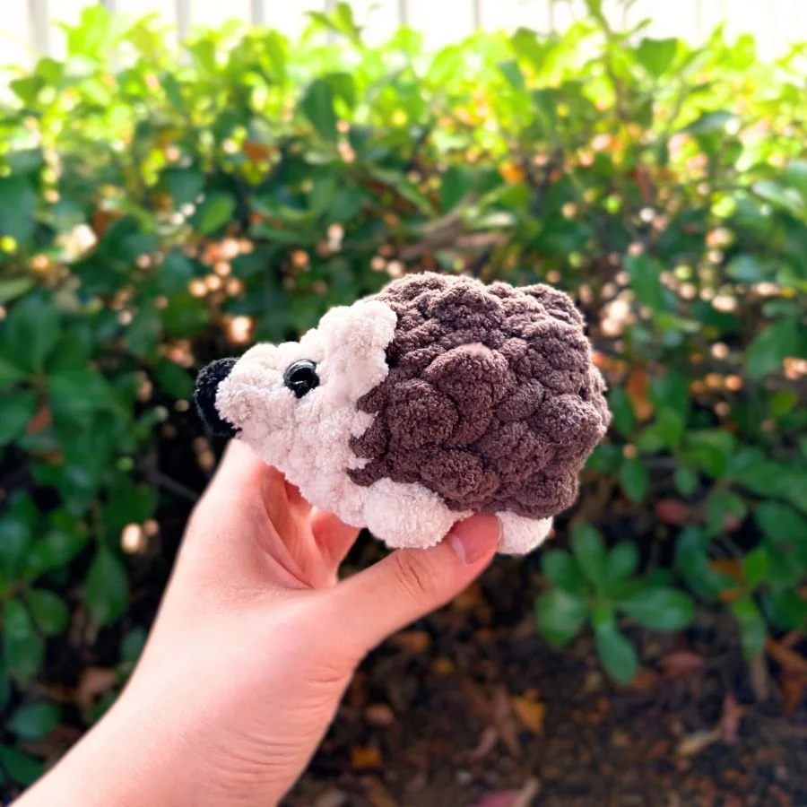 tiny crochet hedgehog made with blanket yarn held by hand in front of green leafy background - free crochet pattern