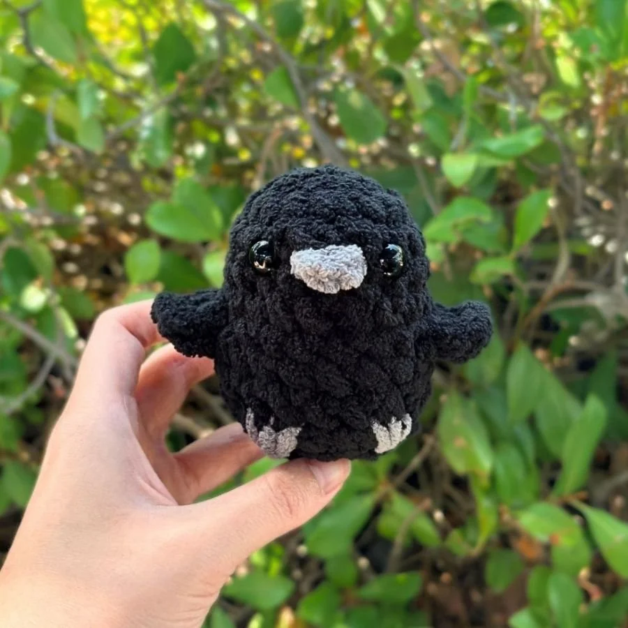 front view of a little crochet no-sew raven made with black and gray yarn held by a hand - free crochet amigurumi raven pattern