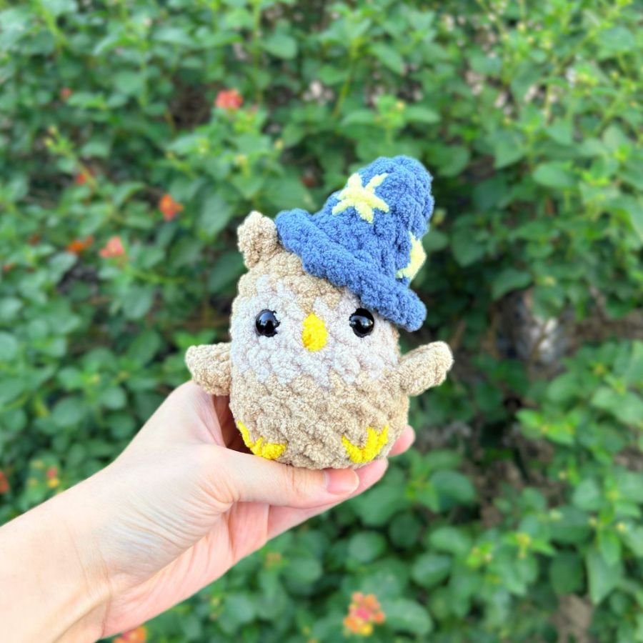 little crochet amigurumi owl held by a hand with a tiny crochet blue wizard hat with yellow embroidered stars - free crochet pattern