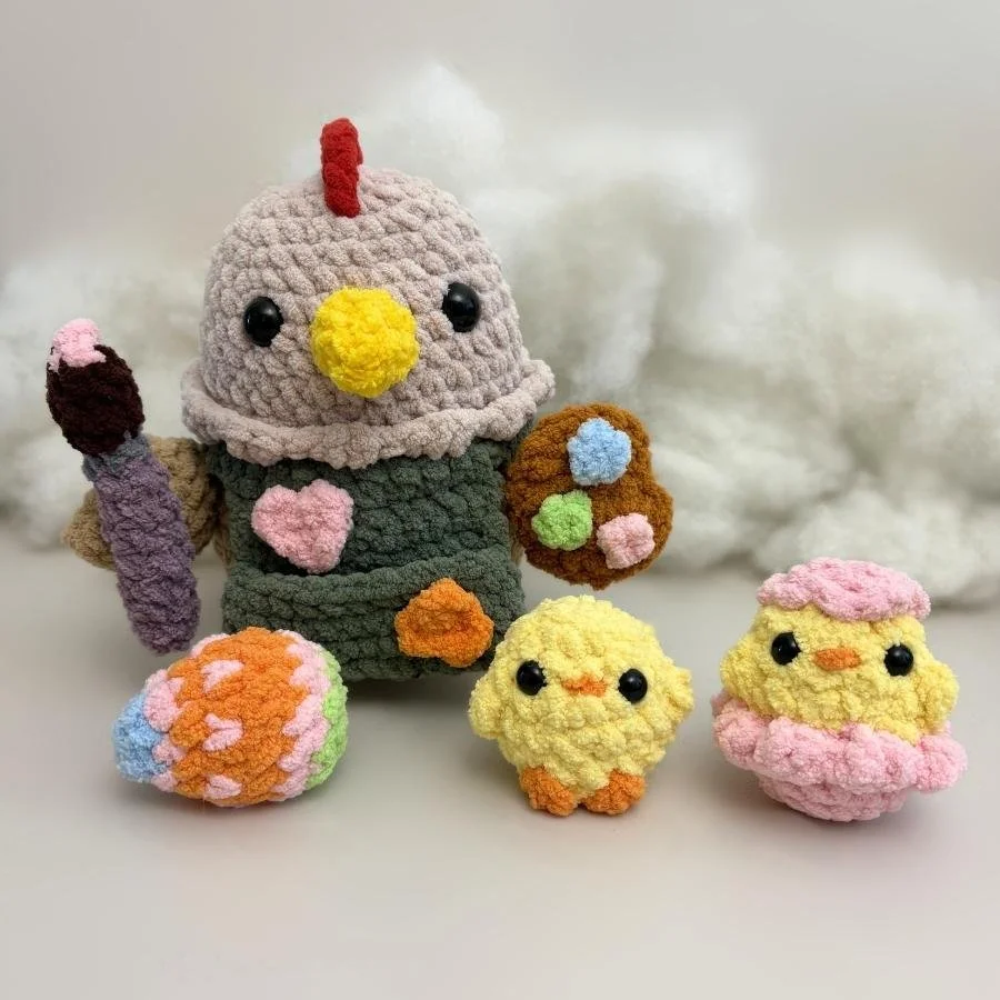 crochet chicken with a paintbrush, paint palette, and wearing a green apron with paint splatters along with an easter egg and a little yellow crochet chick and a crochet hatchling chick in a pink eggshell