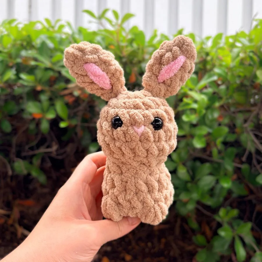 little, brown crochet rabbit with pink ear accents and bobbles for feet held by hand against green bush background - free crochet pattern