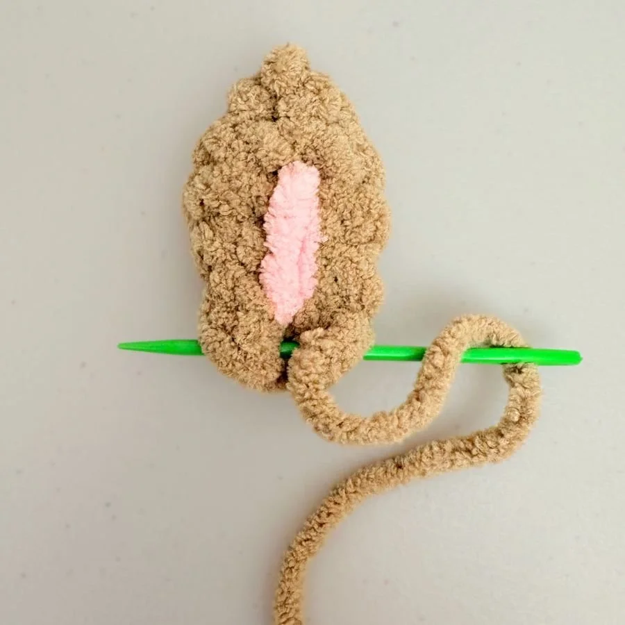 a green needle sewing across the bottom of a brown, crochet rabbit ear to show how to shape the ear