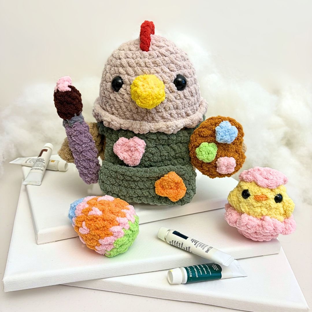 crochet painting chicken wearing a green painter's apron with paint splotches and holding a purple crochet paintbrush and a mini crochet paint palette. Next to it is a crochet easter egg and a crochet hatchling chick with a pink egg shell.
