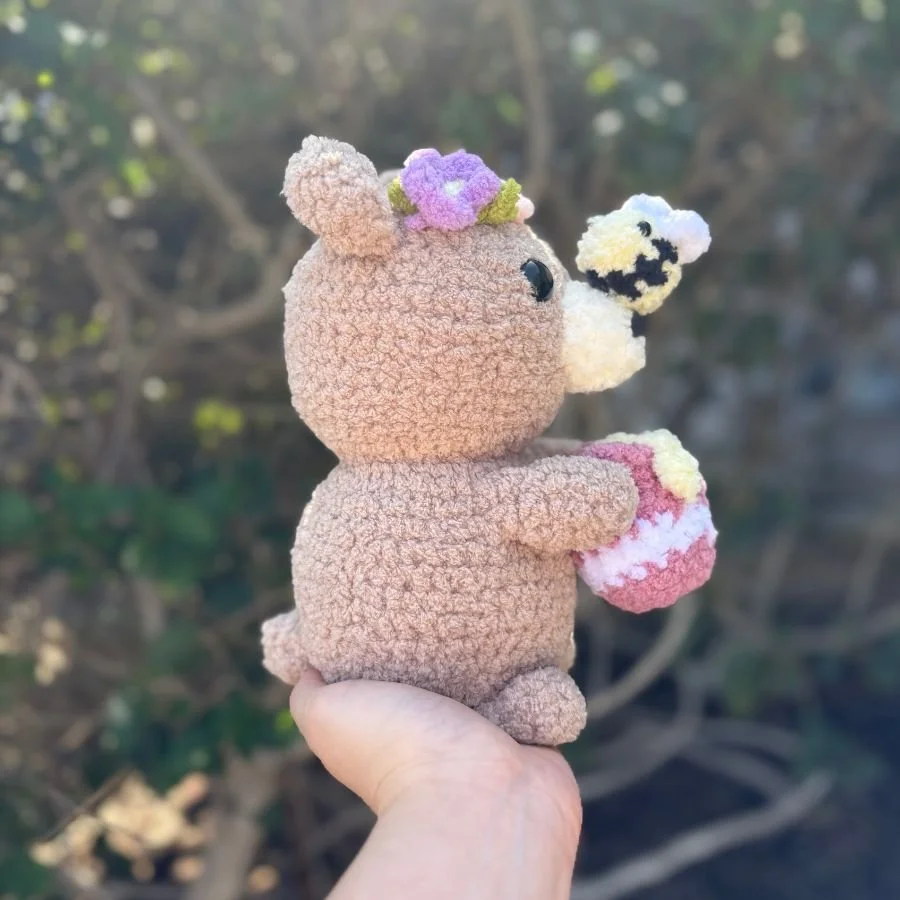 side view of a brown crochet bear with a bee on its nose and holding a honey pot