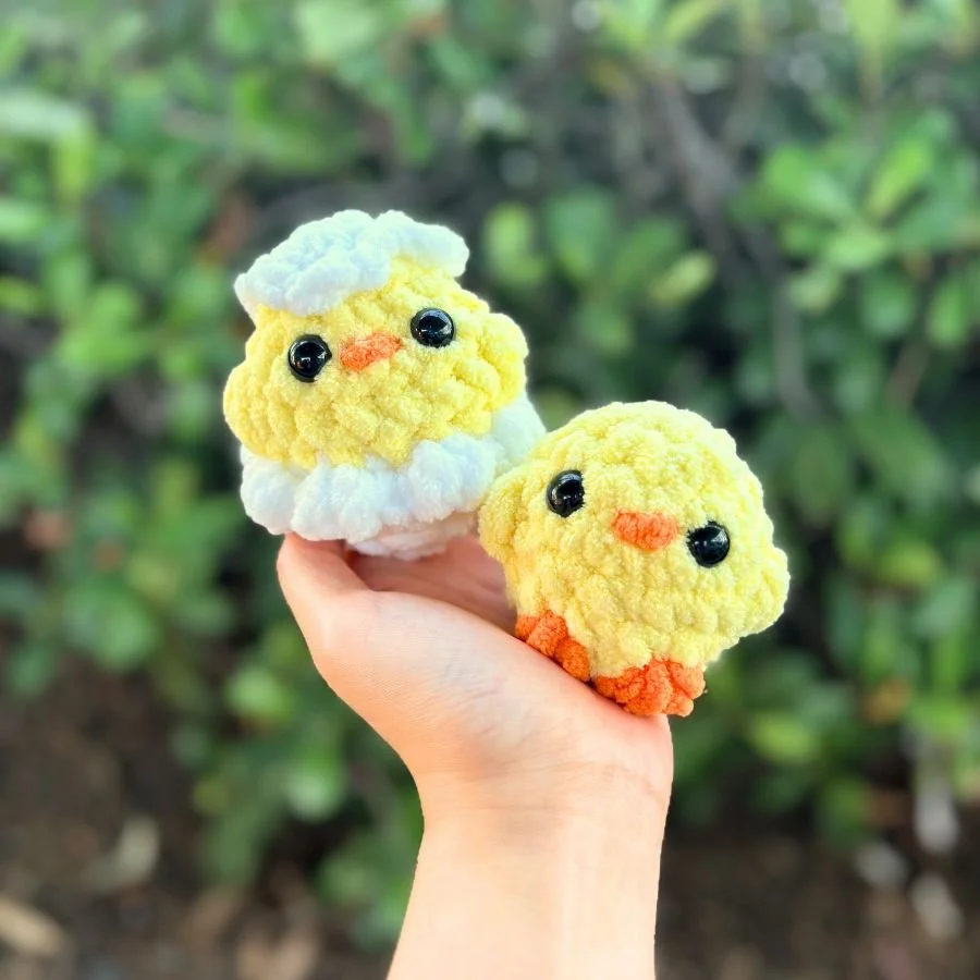 crochet yellow hatchling chick sitting in a white egg shell and an eggshell on its head. Next to it is a crochet yellow chick with an orange beak and feet. They are both being held up in one hand. Free crochet chick pattern.