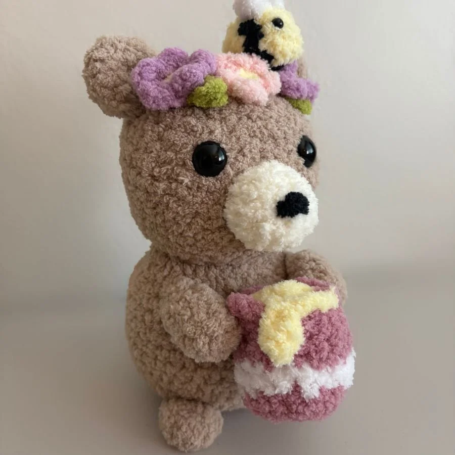 fuzzy brown crochet honey bear with beige snout, a pink and purple flower crown and bee on its head, and holding a honey pot with dripping honey - free crochet pattern honey bear