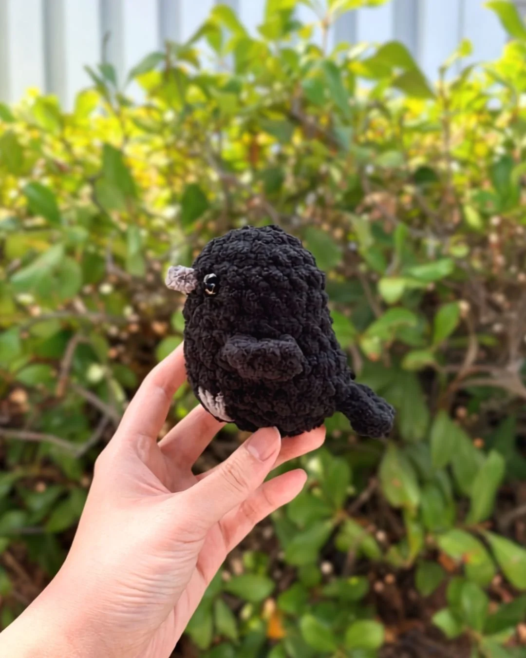little crochet raven made with black blanket yarn and a gray beak and feet, held by a hand in front of a leafy background. Free crochet  no-sew raven pattern