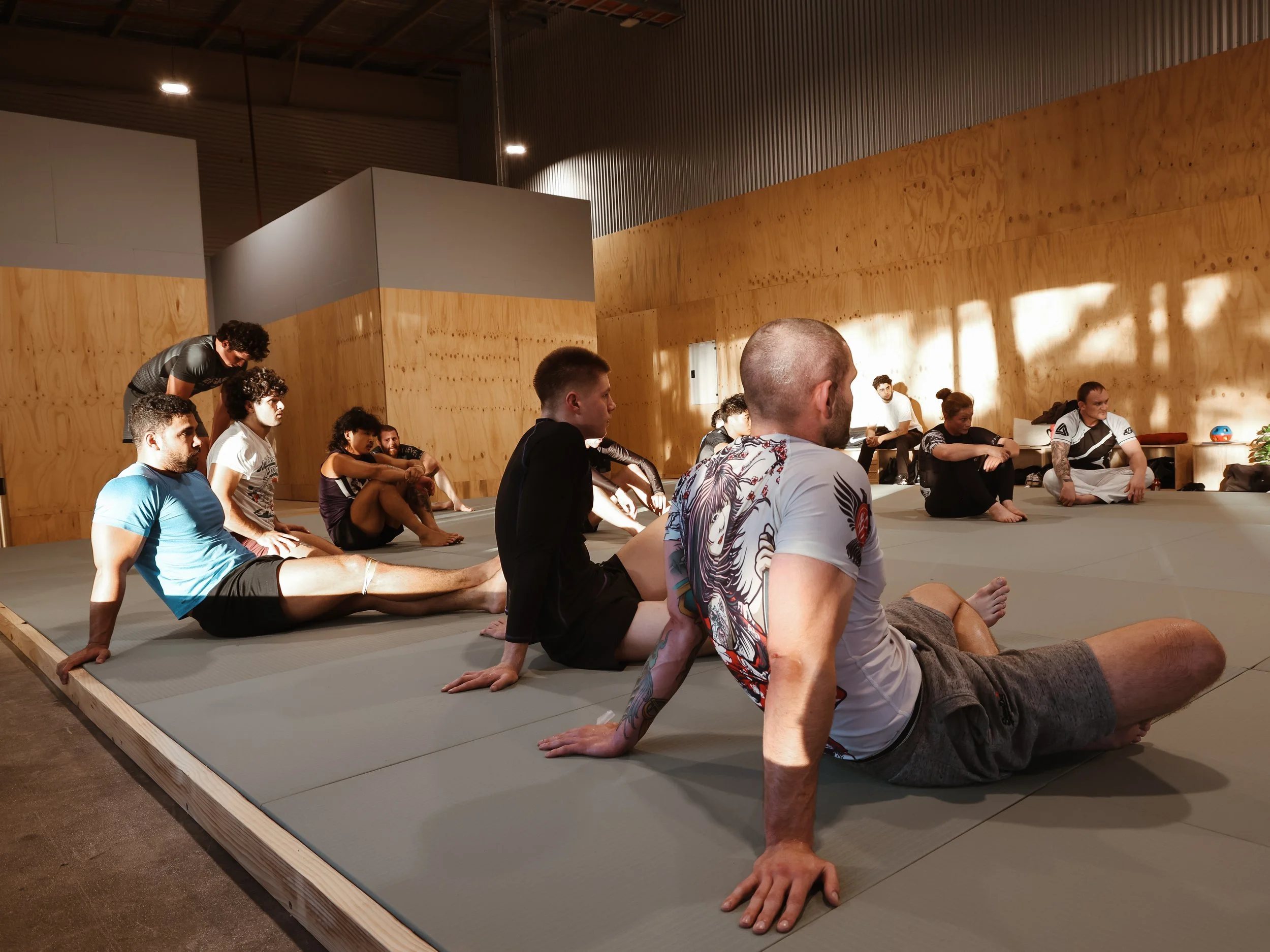 Group of people in a martial arts class practicing stretching on mats in a training facility with wooden walls.