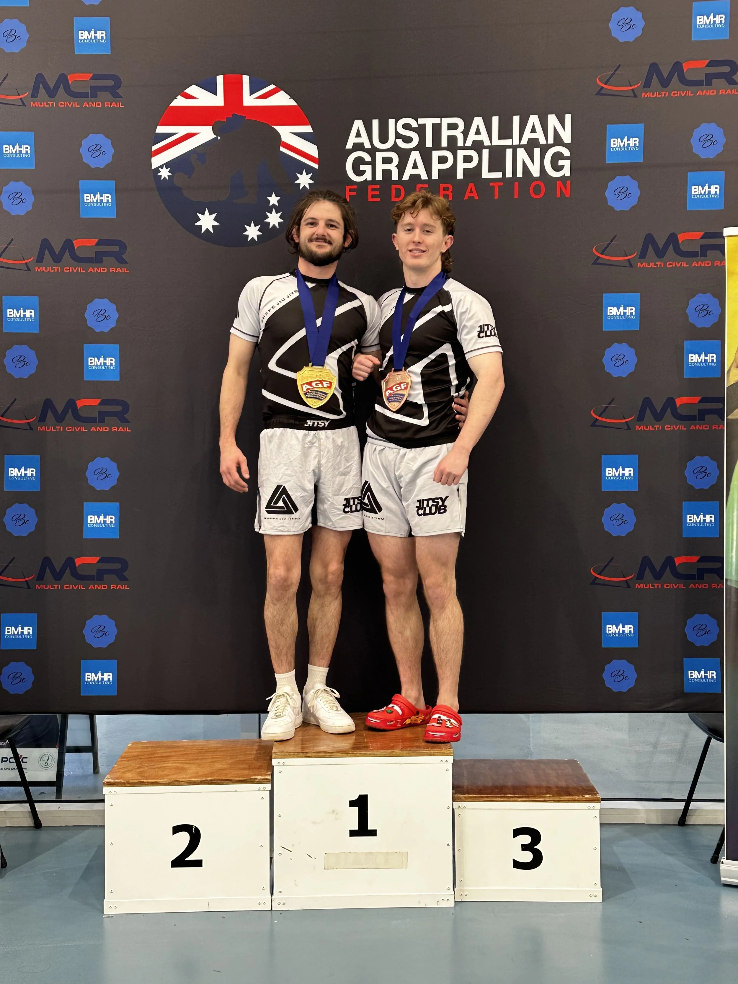 Two male athletes on a winner's podium at an Australian grappling competition. They are smiling, wearing medals, and athletic uniforms, with one in first place and the other in second. A backdrop features the Australian Grappling Federation logo and sponsors.