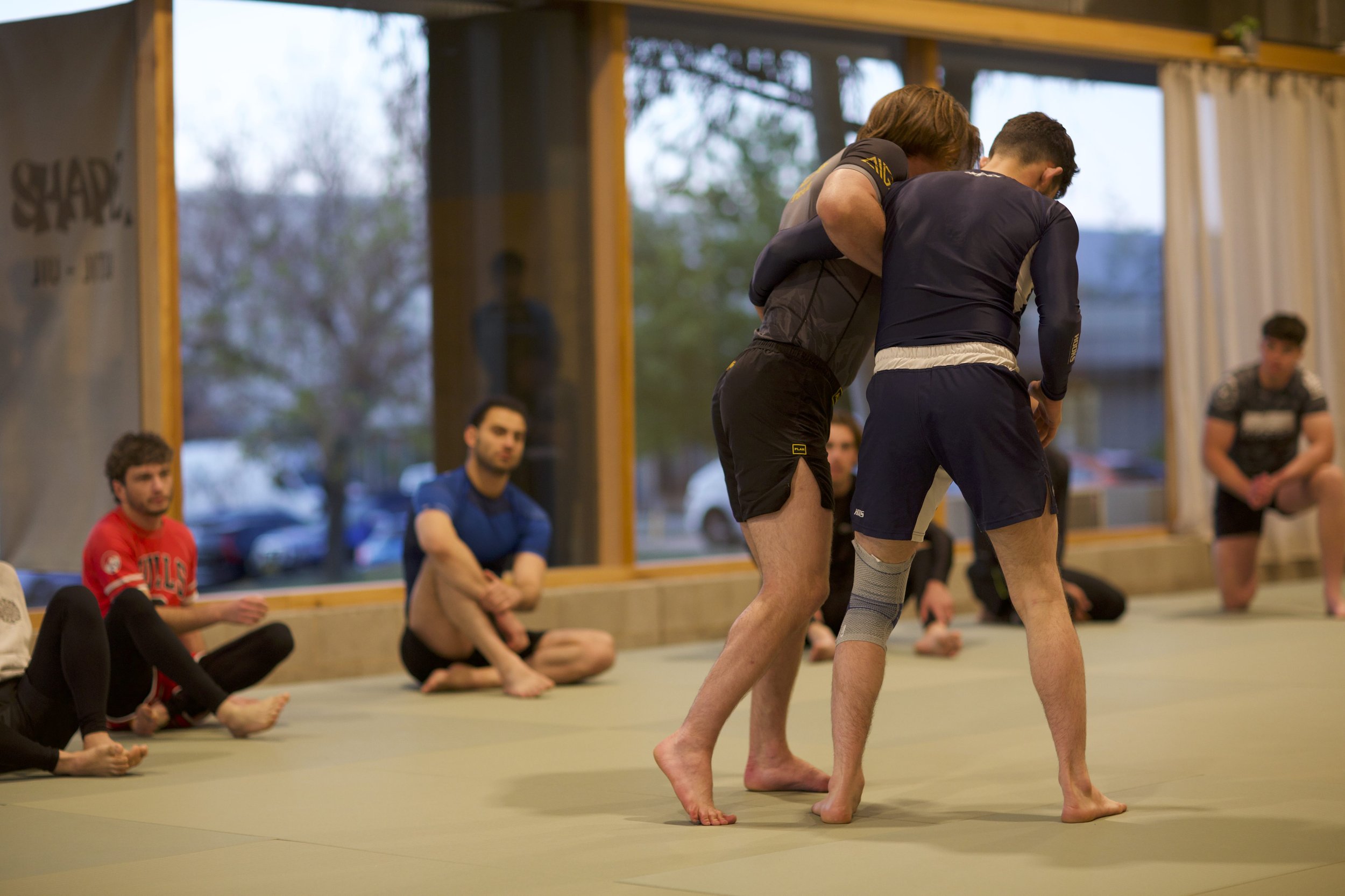 Two men practicing Brazilian Jiu-Jitsu on a mat while others watch, inside a gym with large windows and a few cars outside.