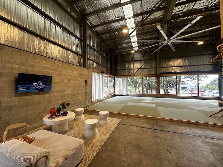 Spacious industrial-style room with concrete floor, metal ceiling, large windows, and a tatami mat area. Contains a sofa, small tables, a wall-mounted TV, and decorative plants.