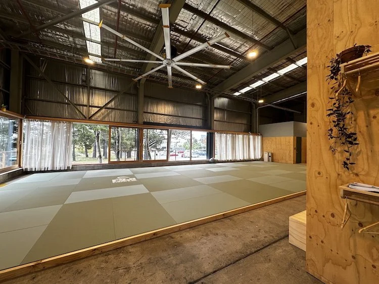 An empty martial arts dojo with tatami mats, large windows with curtains, and industrial ceiling with a large ceiling fan.