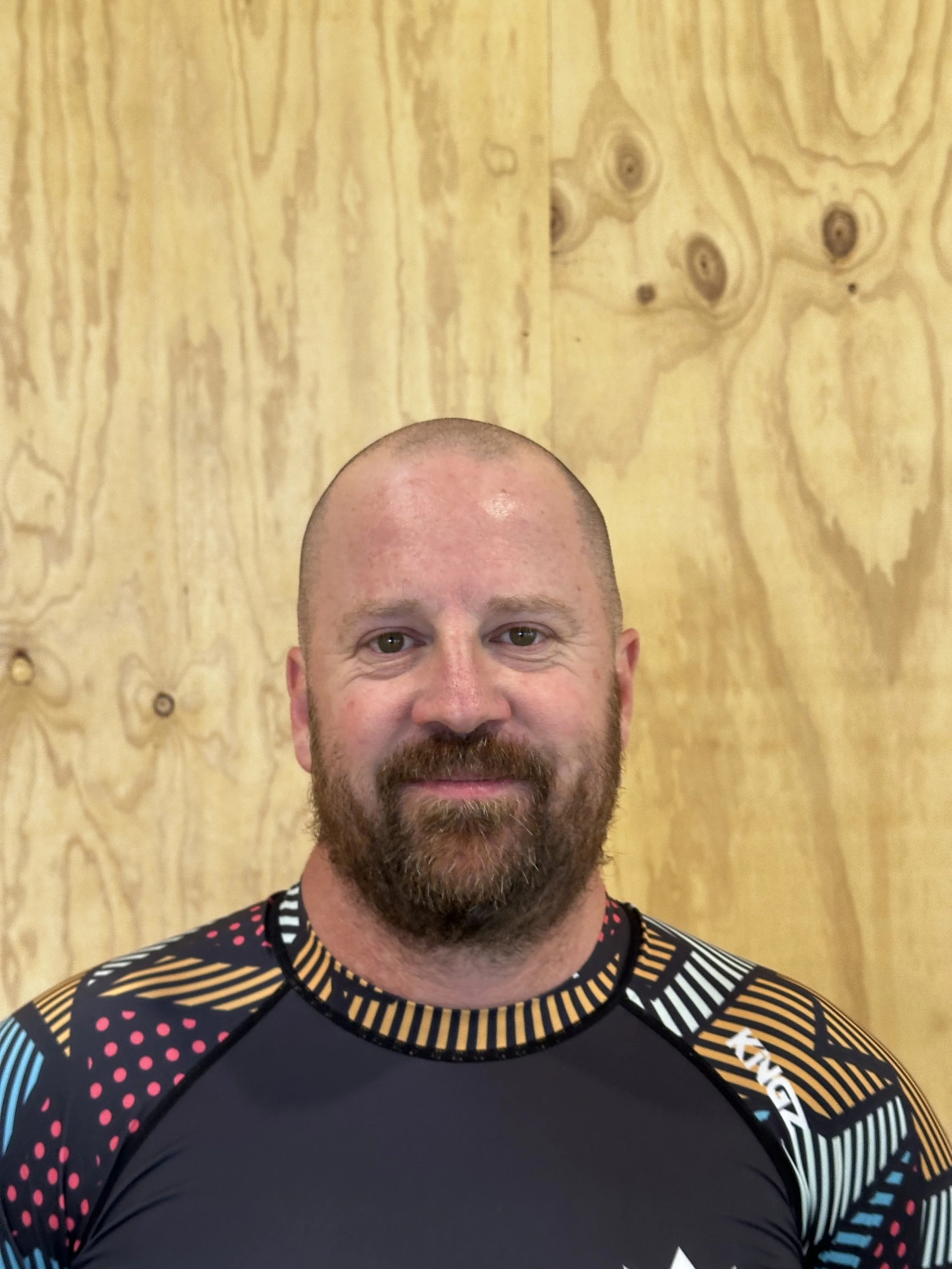 A man with a beard and buzz cut, wearing a black, colorful patterned athletic shirt, standing in front of a wooden background.