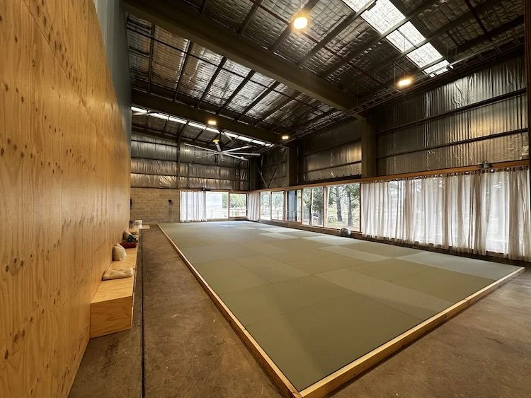 Indoor gym with a matted floor, large windows covered with curtains, and a high ceiling with lights and a fan.