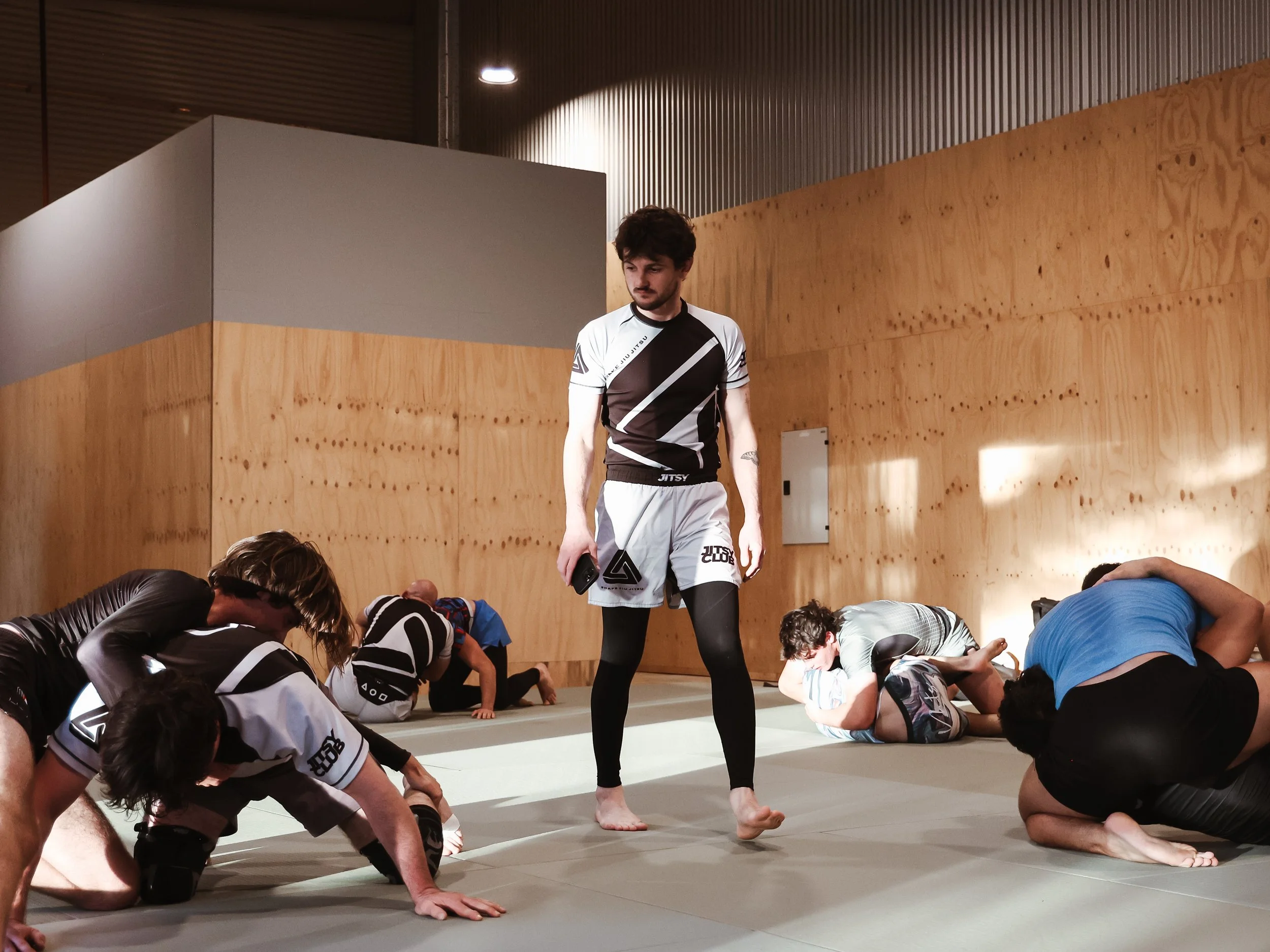 A martial arts instructor stands on a mat while students practice grappling techniques on the ground in a dojo with wooden and gray walls.